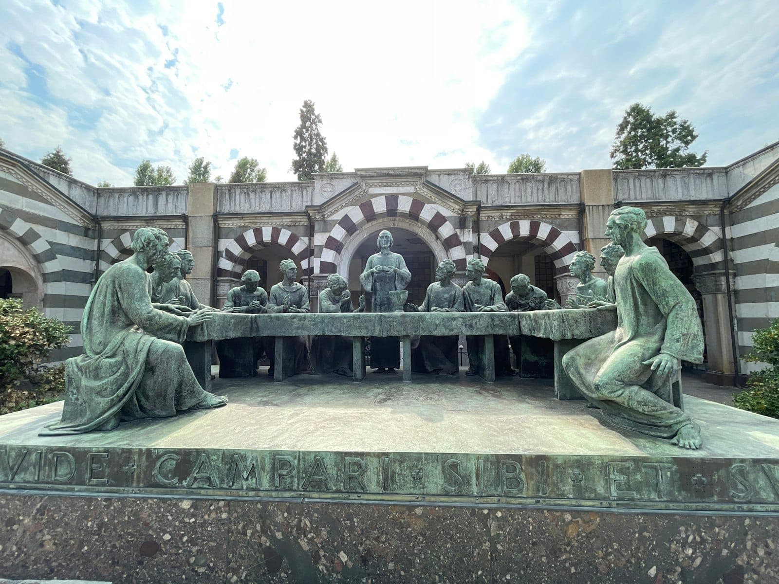Scultura in bronzo dell'Ultima Cena nel cortile del Cimitero Monumentale di Milano, con figure dettagliate e archi a strisce visibili sotto un cielo parzialmente nuvoloso.