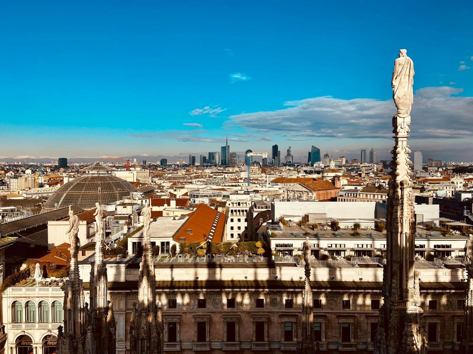 Panoramic rooftop view from Milan's Duomo with Gothic spires in the foreground and modern skyscrapers of the city skyline under a bright blue sky.
