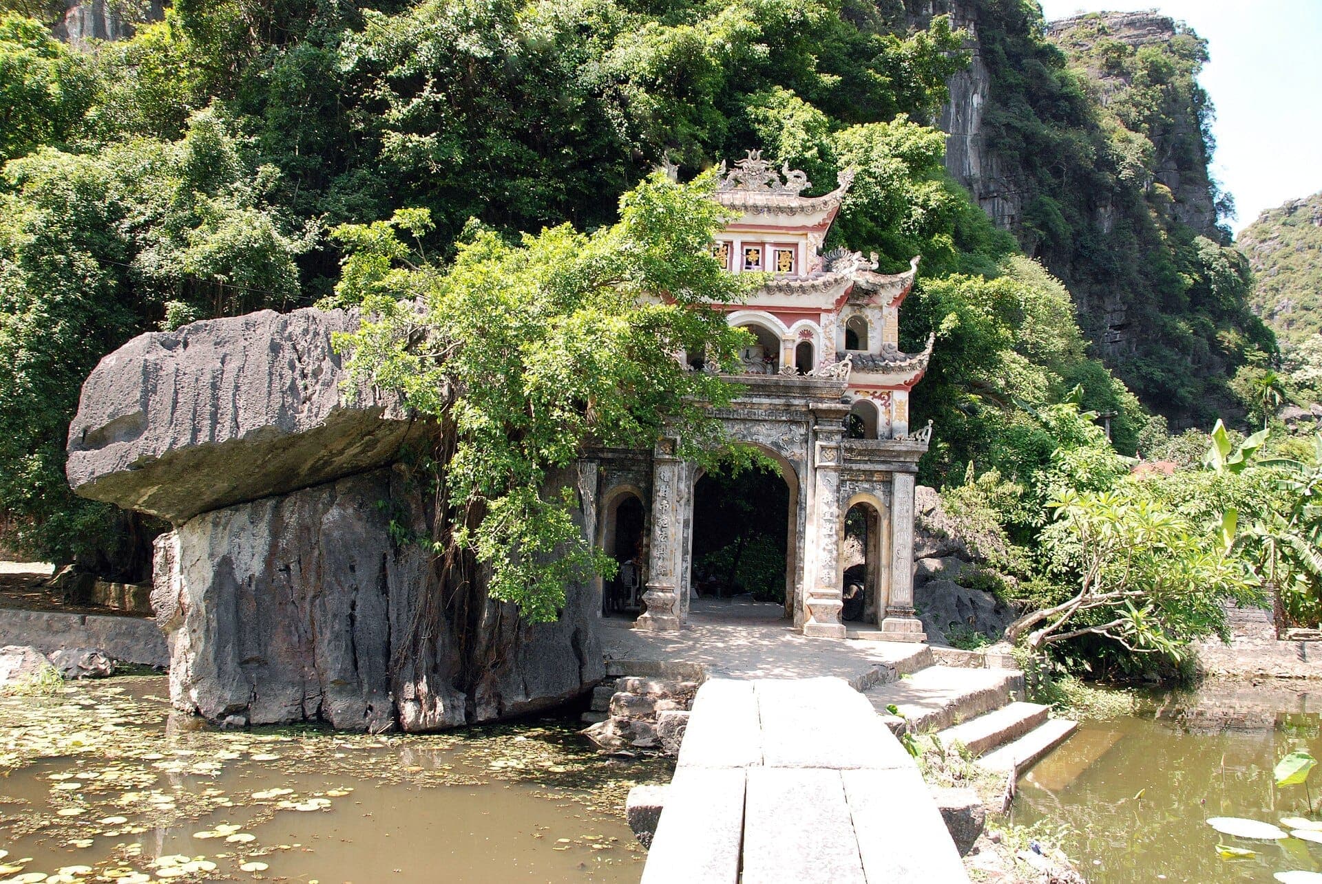 Lối vào Chùa Bích Động được bao quanh bởi cây xanh tươi tốt, vách đá vôi và hồ sen dưới ánh sáng ban ngày.