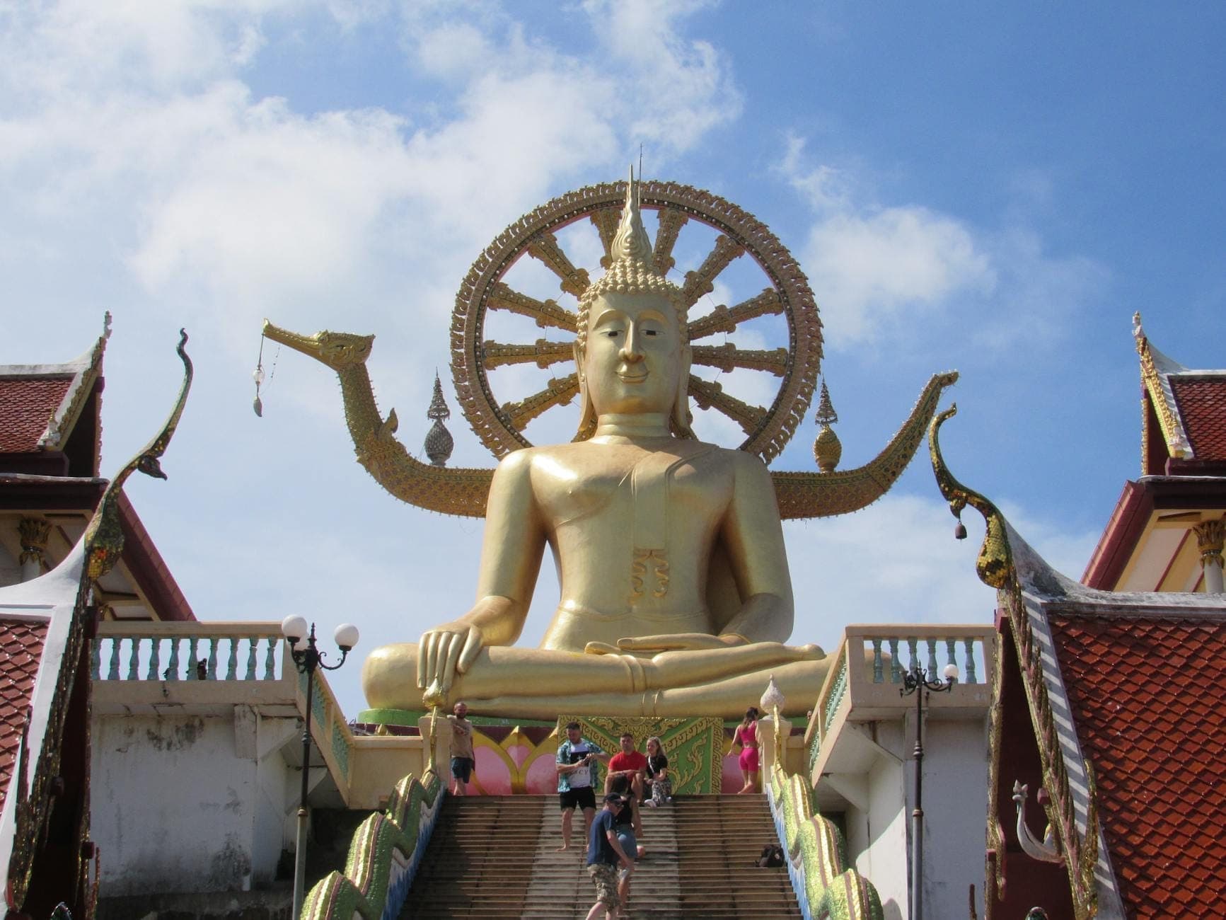 Large golden Buddha statue at the top of an open stairway under blue sky, with temple rooftops and visitors ascending the steps.