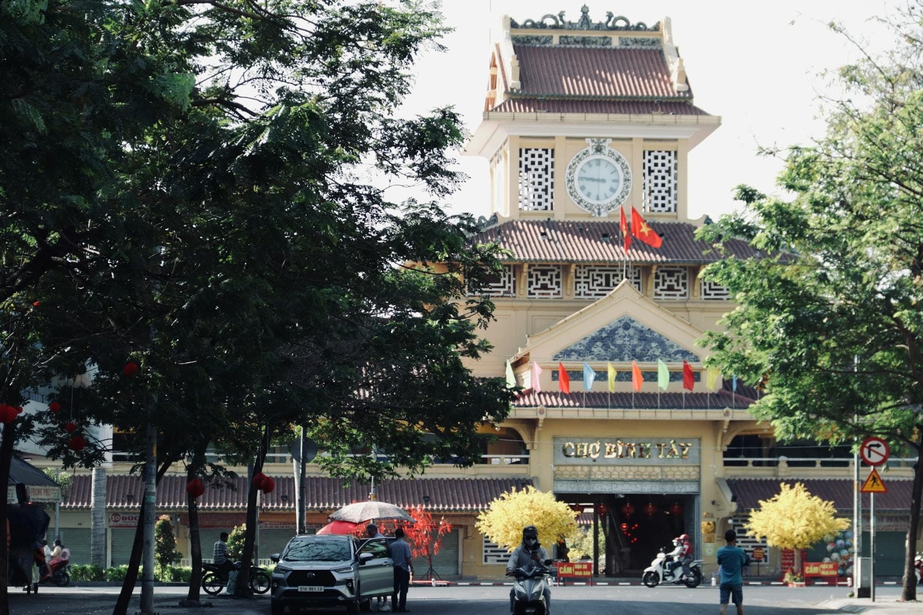 Mặt tiền Chợ Bình Tây với tường vàng ochre đặc trưng, tháp đồng hồ trung tâm, cờ phướn và hàng cây xanh tôn vinh lối vào trong một ngày nắng đẹp.