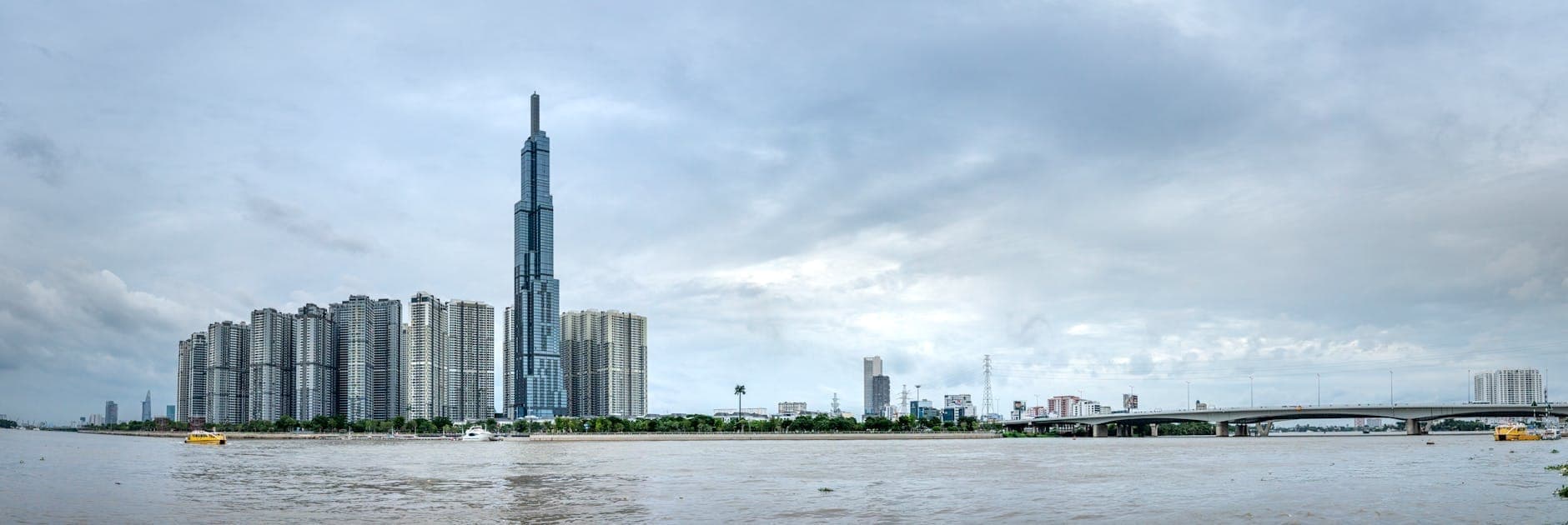 Panoramikong tanawin ng distrito ng Bình Thạnh na may mga modernong mataas na gusali at Landmark 81 sa tabi ng Ilog Saigon sa ilalim ng ulap na langit.