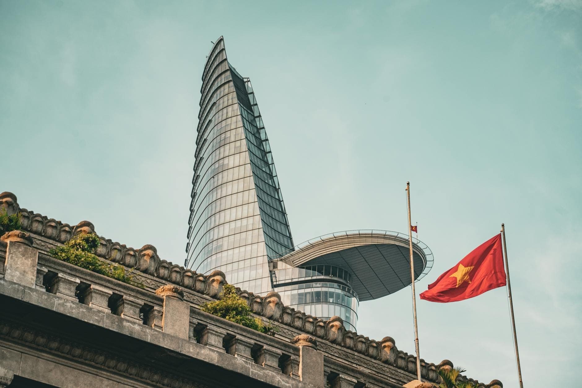 Tòa nhà Bitexco Financial Tower vươn lên trên một tòa nhà cũ có cắm cờ Việt Nam, nổi bật trên nền trời xanh trong khu trung tâm Thành phố Hồ Chí Minh.