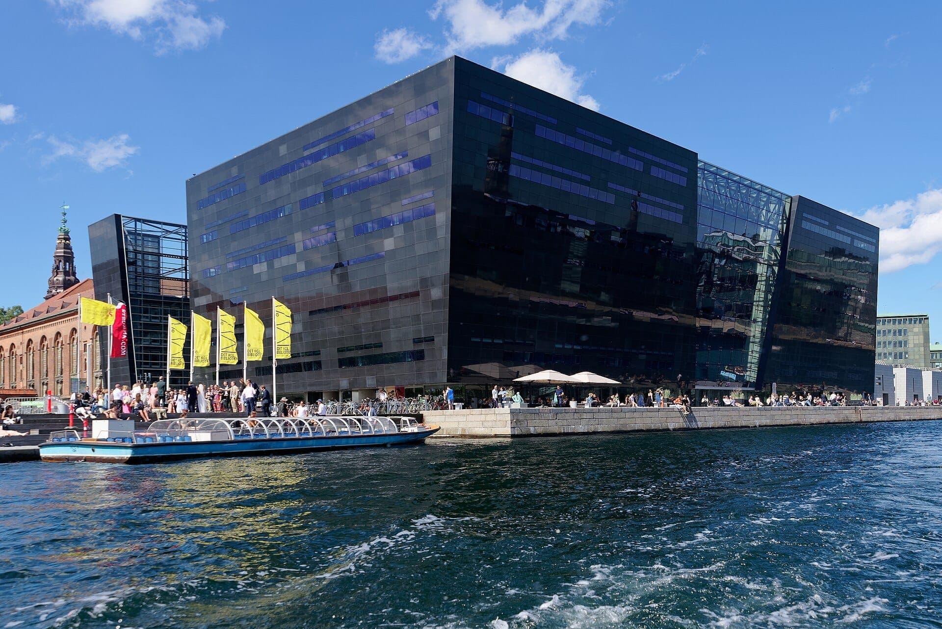 Modernes Black Diamond Bibliotheksgebäude mit spiegelnder schwarzer Fassade, Menschen an der Uferpromenade, gelben Flaggen und einem Kanalboot unter einem klaren blauen Himmel in Kopenhagen.