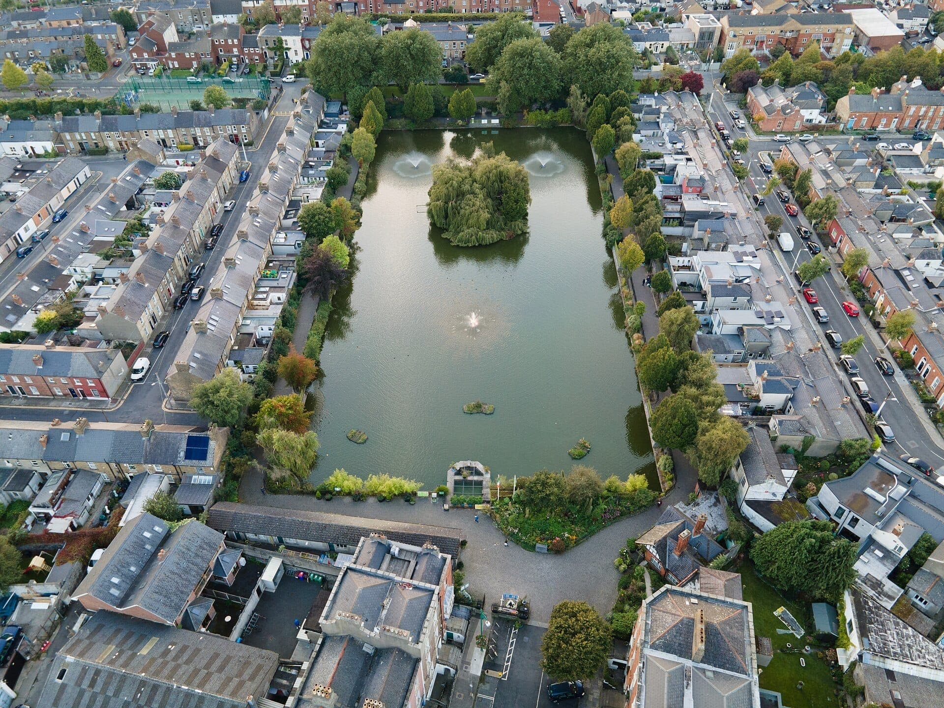 Vista aérea del Blessington Street Basin en Dublín, con el lago central, fuentes, senderos bordeados de árboles y las calles residenciales circundantes.