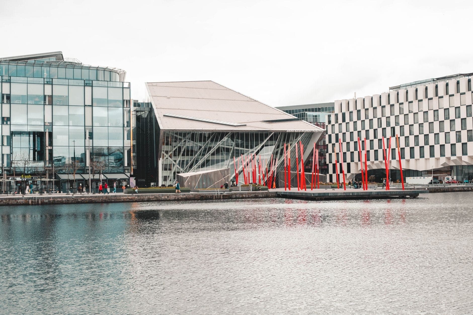 Außenansicht des Bord Gáis Energy Theatre in Dublin mit seiner modernen Architektur und dem markanten geneigten Dach, umgeben von Wasser und den leuchtend roten Masten des Grand Canal Square.