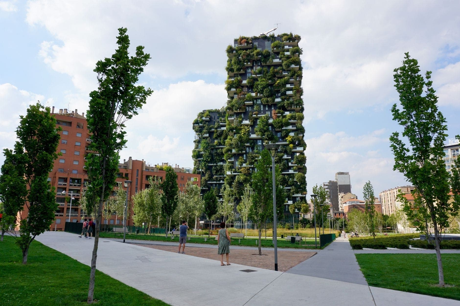 Bosco Verticale’s lush green towers rise behind a landscaped urban park with paths, young trees, and people walking under a bright, partly cloudy sky.