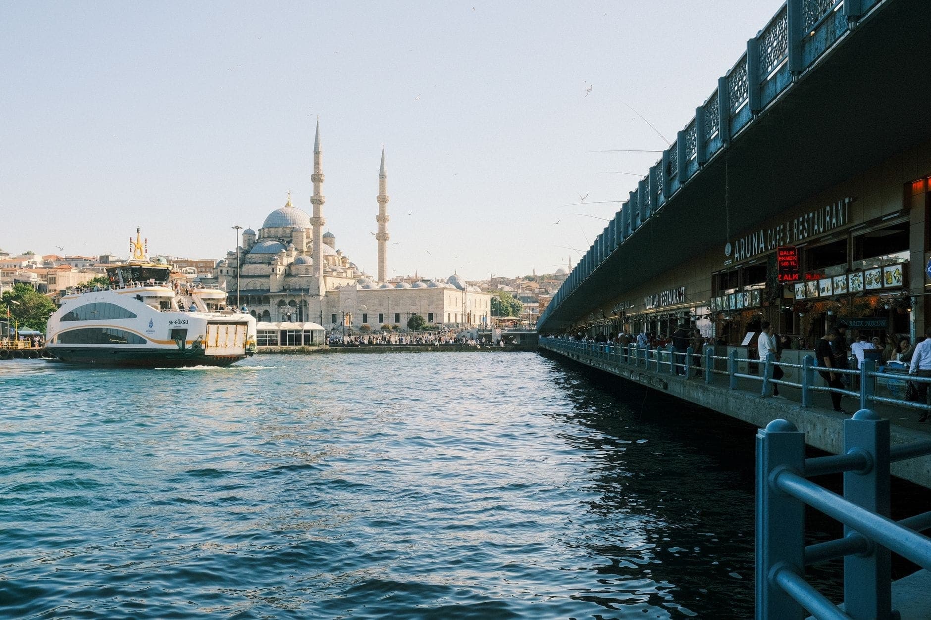 Widok na molo Eminönü i Most Galata nad Bosforem z promem i widocznym Nowym Meczetem, ludzie spacerujący wzdłuż nabrzeża.