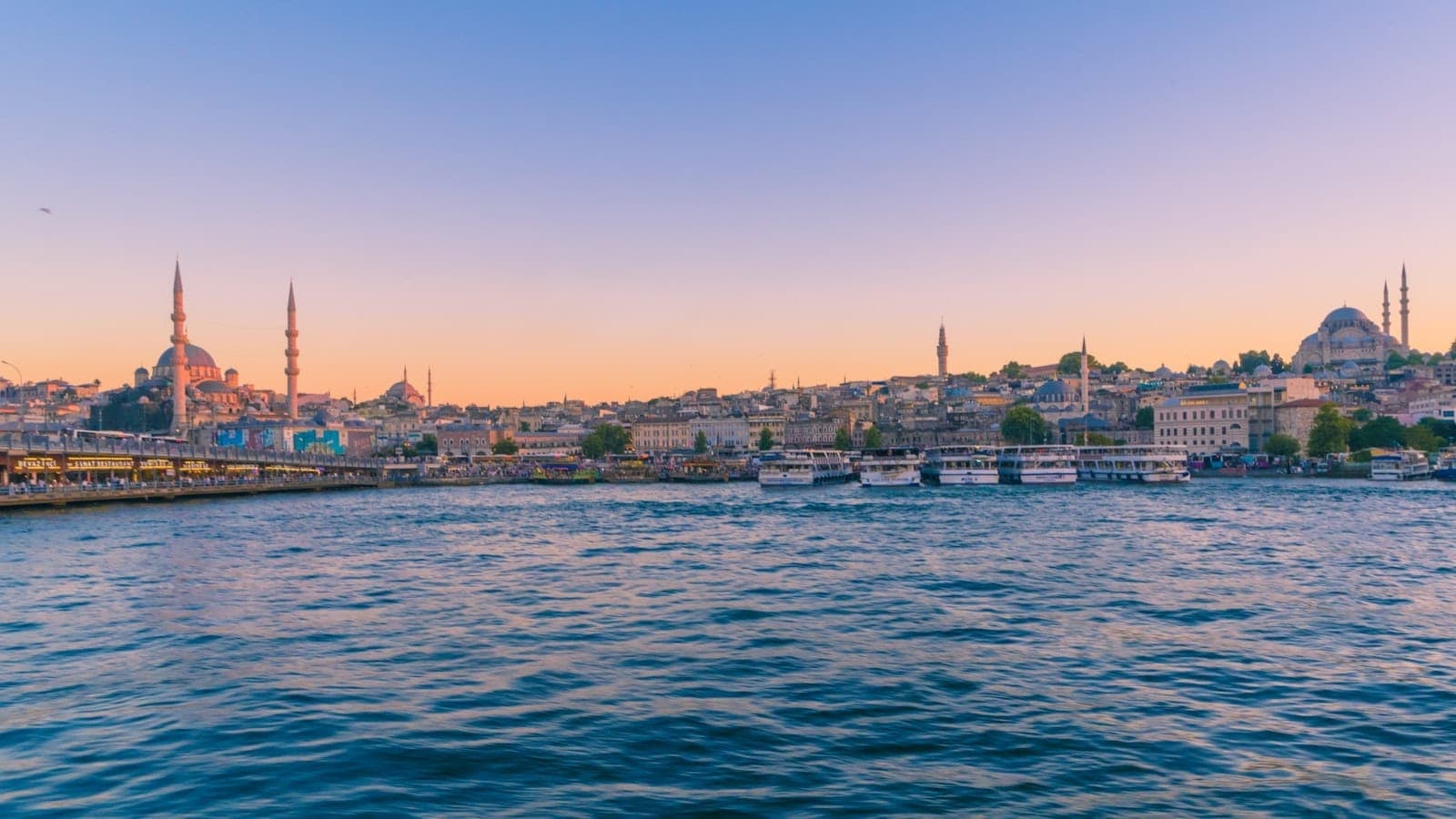 Panoramablick auf Istanbuls Skyline bei Sonnenuntergang mit Moscheen und Minaretten, buntem Himmel, mehreren Fähren und Booten auf dem Bosporus sowie Stadtgebäuden entlang der Uferpromenade.