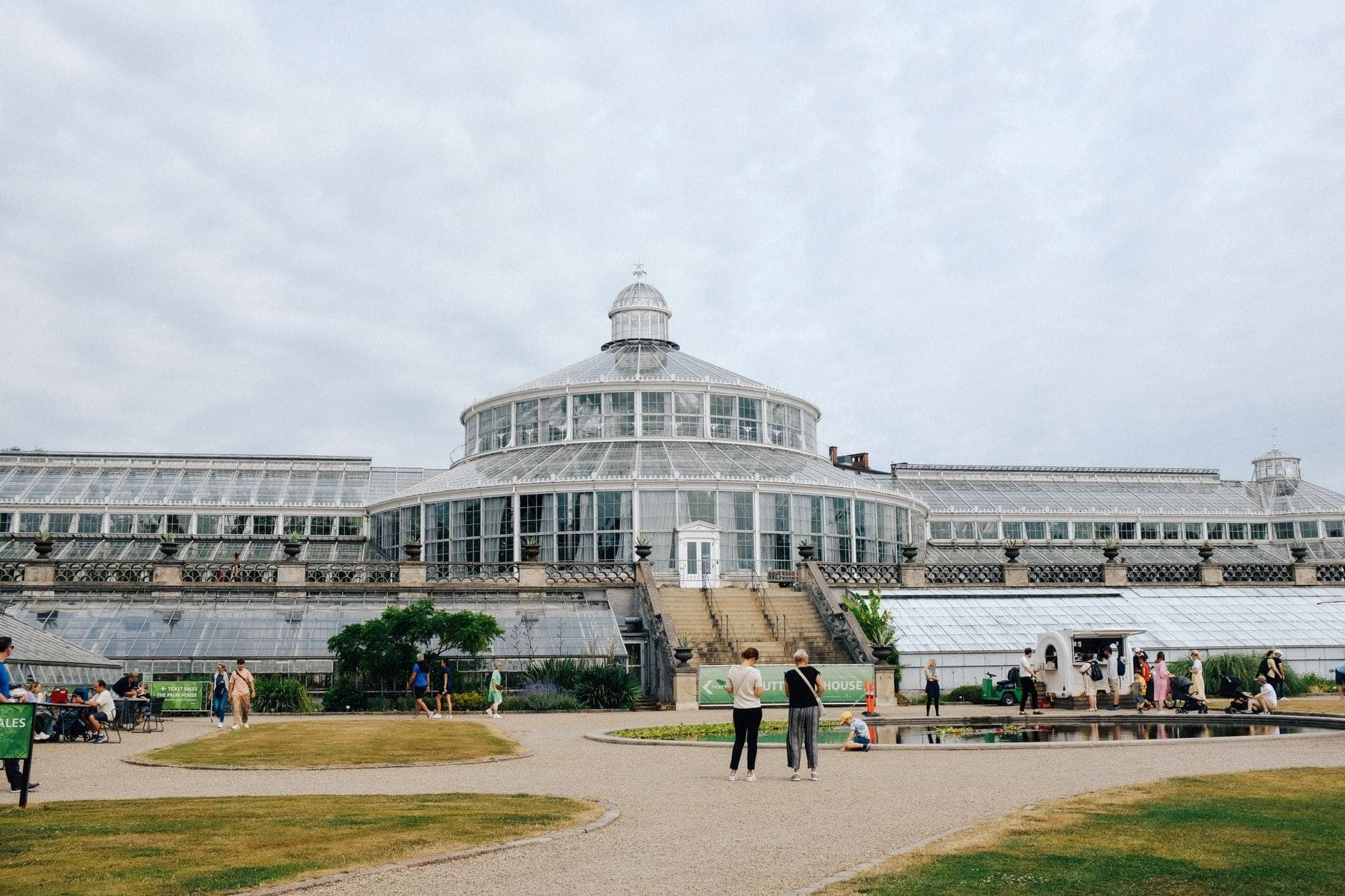 Weitwinkelpanorama des ikonischen viktorianischen Gewächshauses im Botanischen Garten Kopenhagens, mit Menschen, die auf Wegen zwischen Grünpflanzen und offenem Himmel spazieren und entspannen.