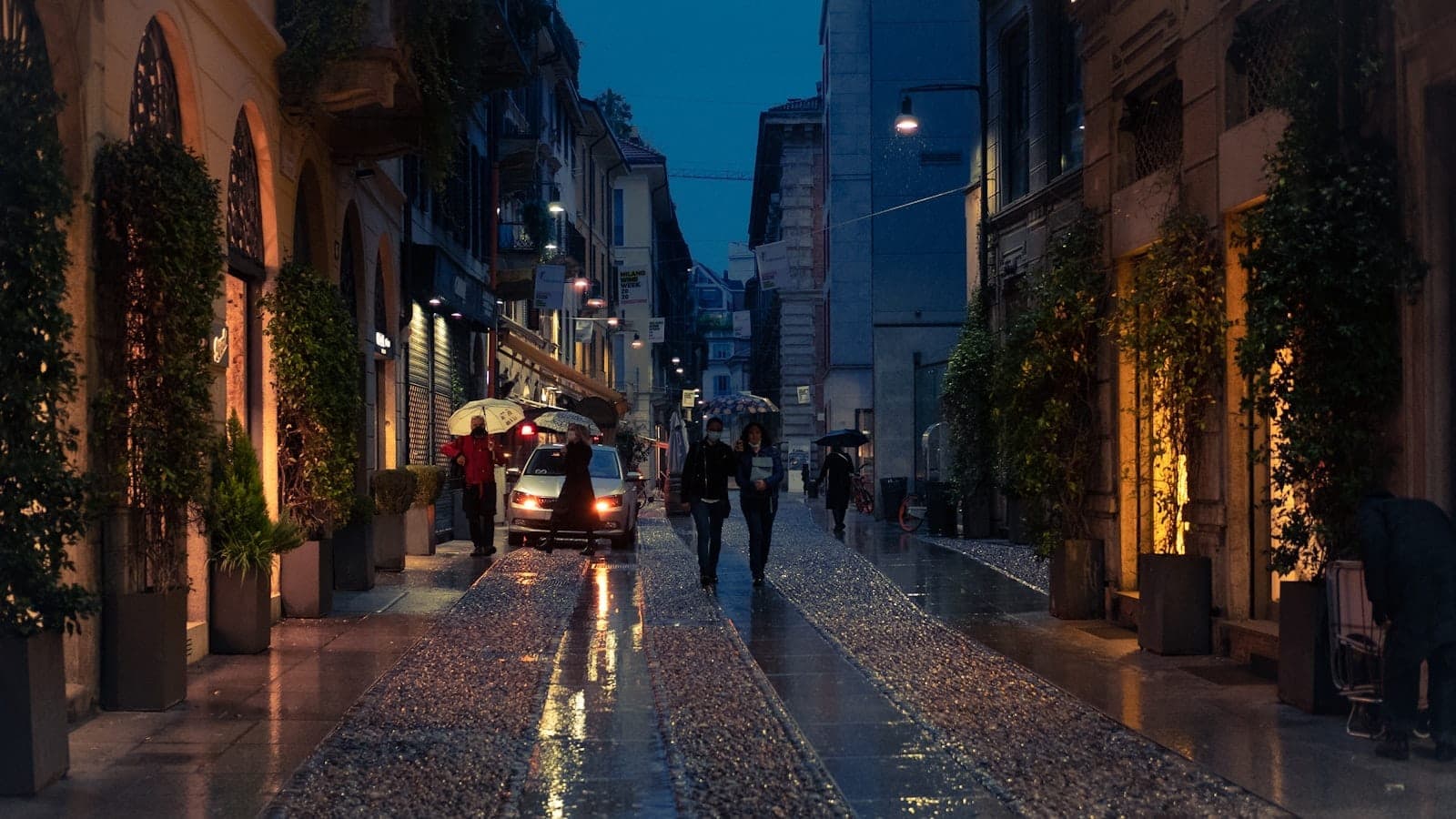 Evening view of a cobbled street in Brera, Milan, lined with boutiques and restaurants, softly lit, with people walking and carrying umbrellas.