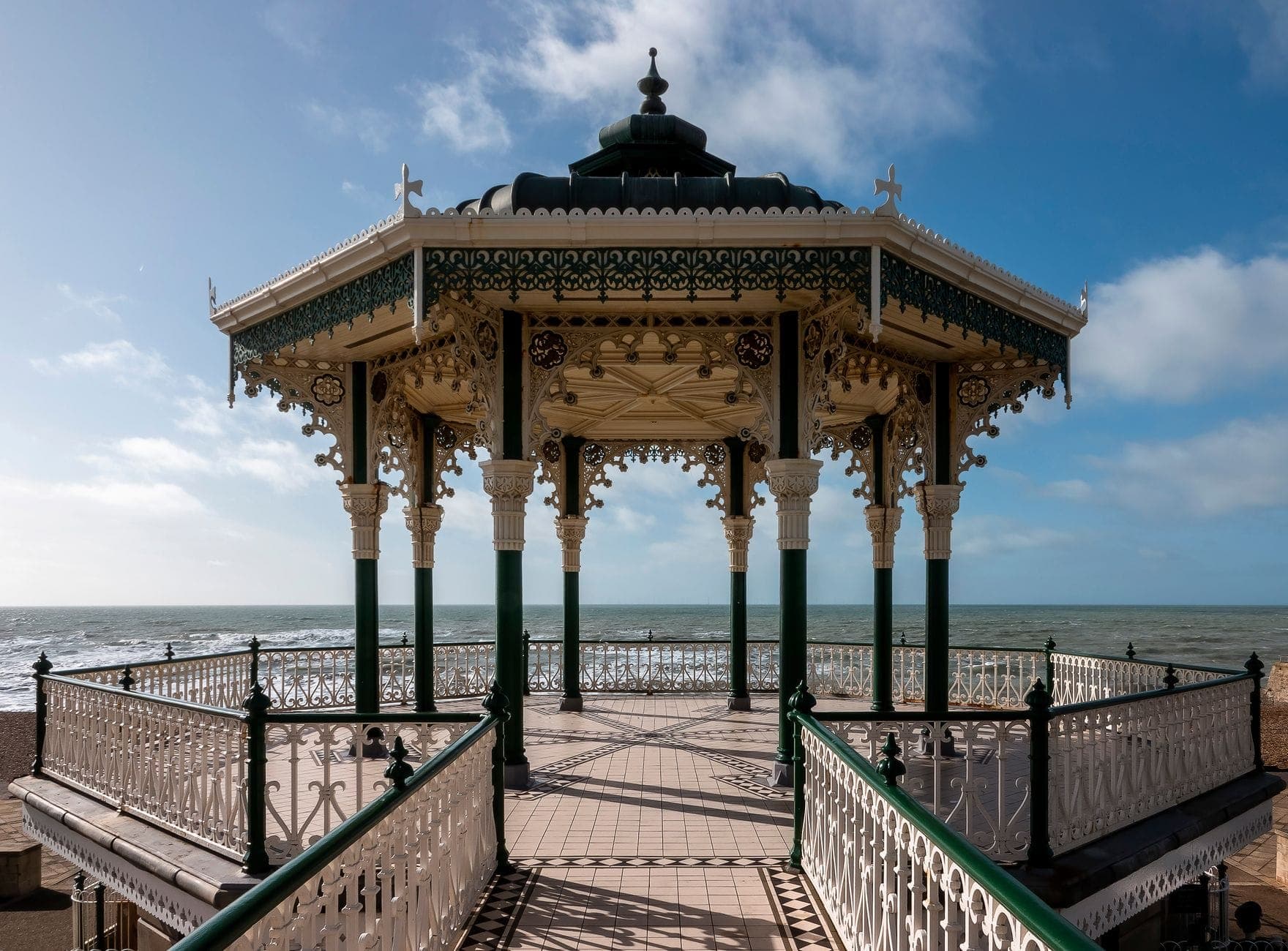 Brighton Bandstand nhìn từ lối đi dạo ven biển, nổi bật với kết cấu sắt đúc cầu kỳ kiểu Victoria, phía sau là biển Anh vào ngày nắng đẹp.