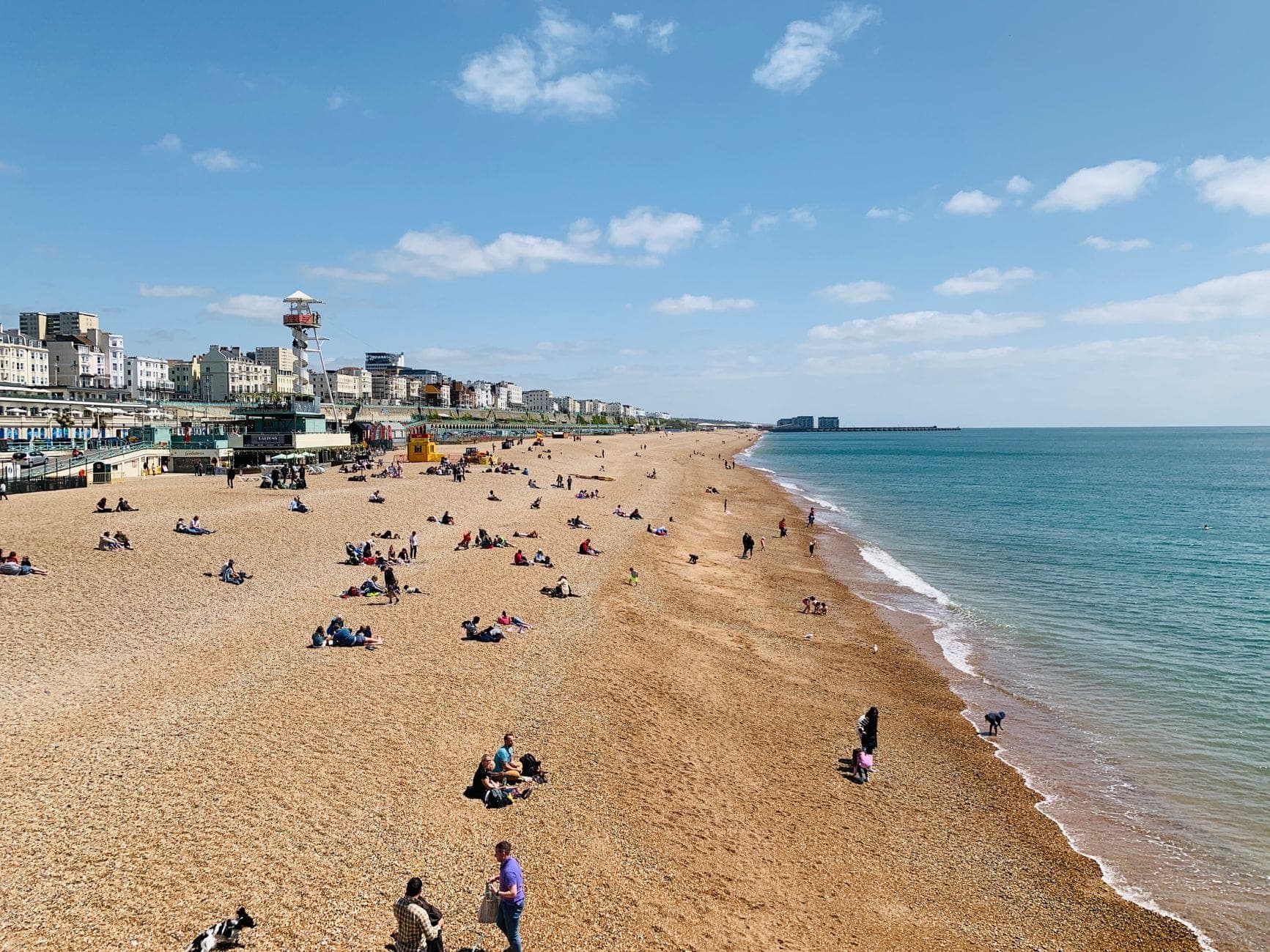 Toàn cảnh bãi biển Brighton đầy sỏi dưới nắng với người dạo chơi trên bờ, các tòa nhà và tháp i360 phía sau.