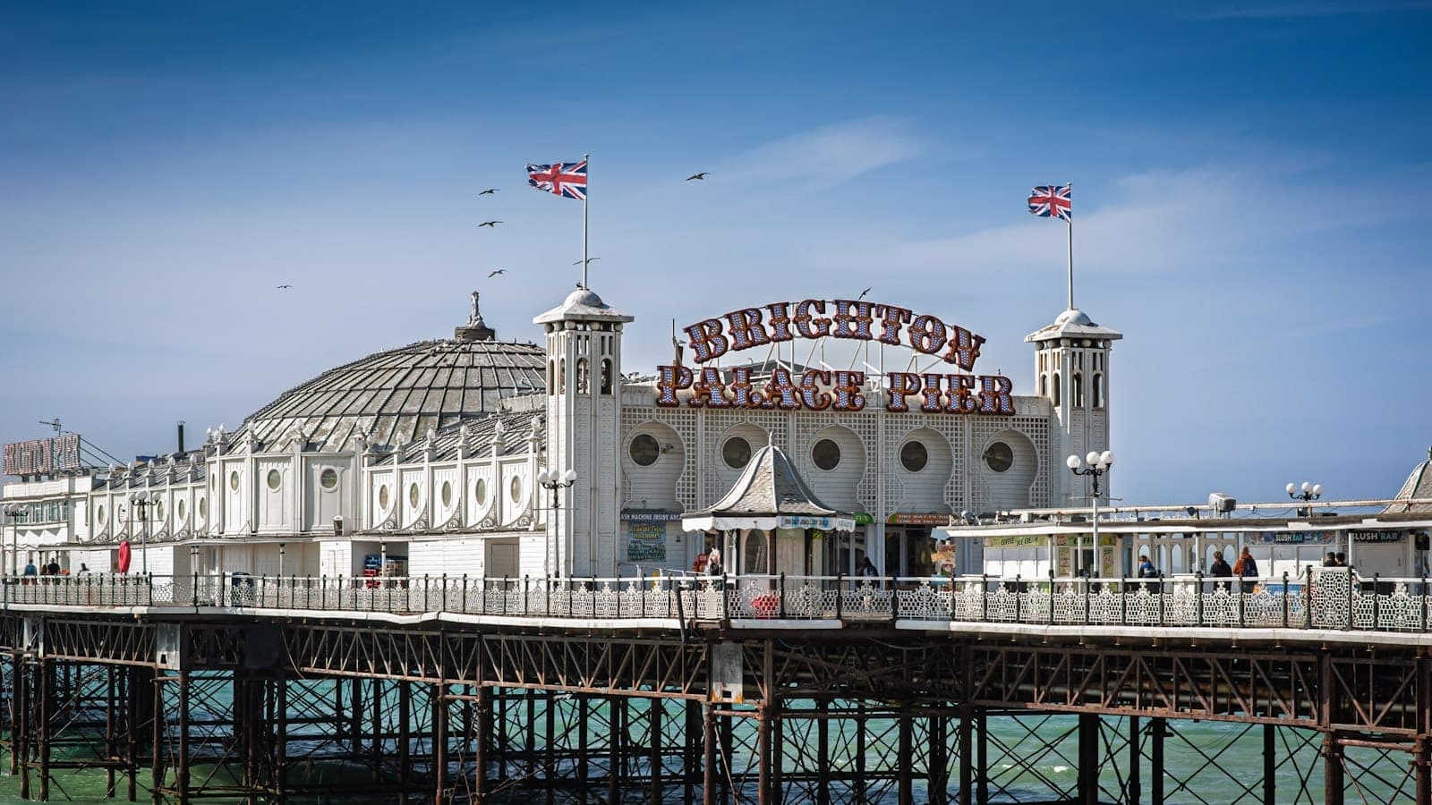 Bến tàu Brighton Palace chụp từ xa dưới bầu trời xanh nổi bật và các chú mòng biển, nổi bật kiến trúc trắng kiểu Victoria cùng không khí nhộn nhịp bên bờ biển.