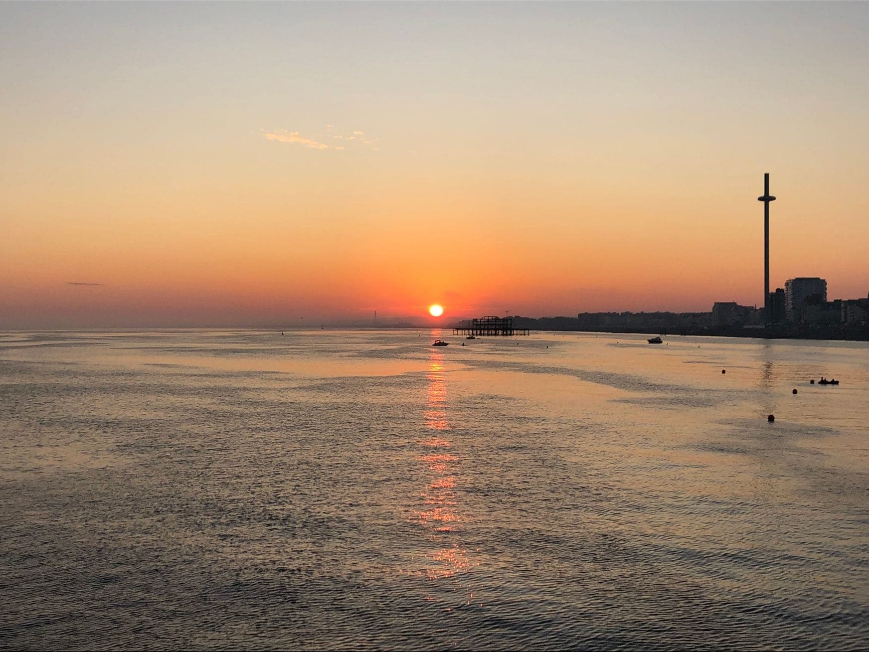 Tháp quan sát Brighton i360 nổi bật trên nền trời hoàng hôn cam, biển êm và cầu tàu phía xa bên bờ biển Brighton.