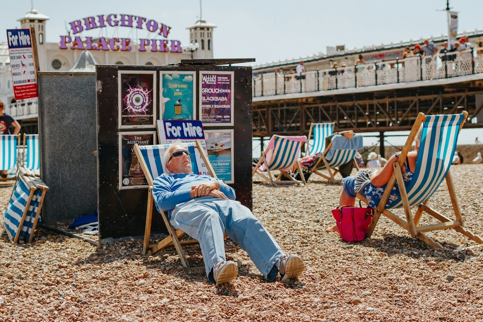 Mọi người thư giãn trên bãi biển Brighton ngồi ghế tựa, phía xa là Cầu tàu Brighton Palace dưới bầu trời hè trong xanh.
