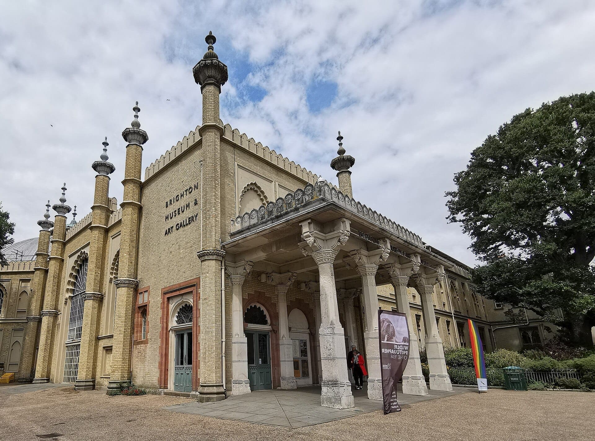 Lối vào trang trí cầu kỳ của Brighton Museum & Art Gallery với các cột Indo-Saracenic, tường gạch màu kem và cửa sổ vòm dưới bầu trời có mây một phần.