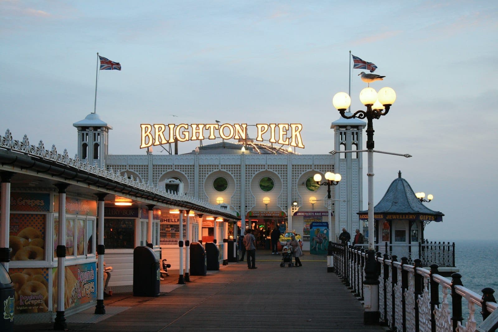 Khung cảnh chiều tà dọc cầu tàu Brighton, người đi dạo dưới ánh đèn lấp lánh và tấm biển biểu tượng, không khí biển cùng bầu trời nhiều mây.