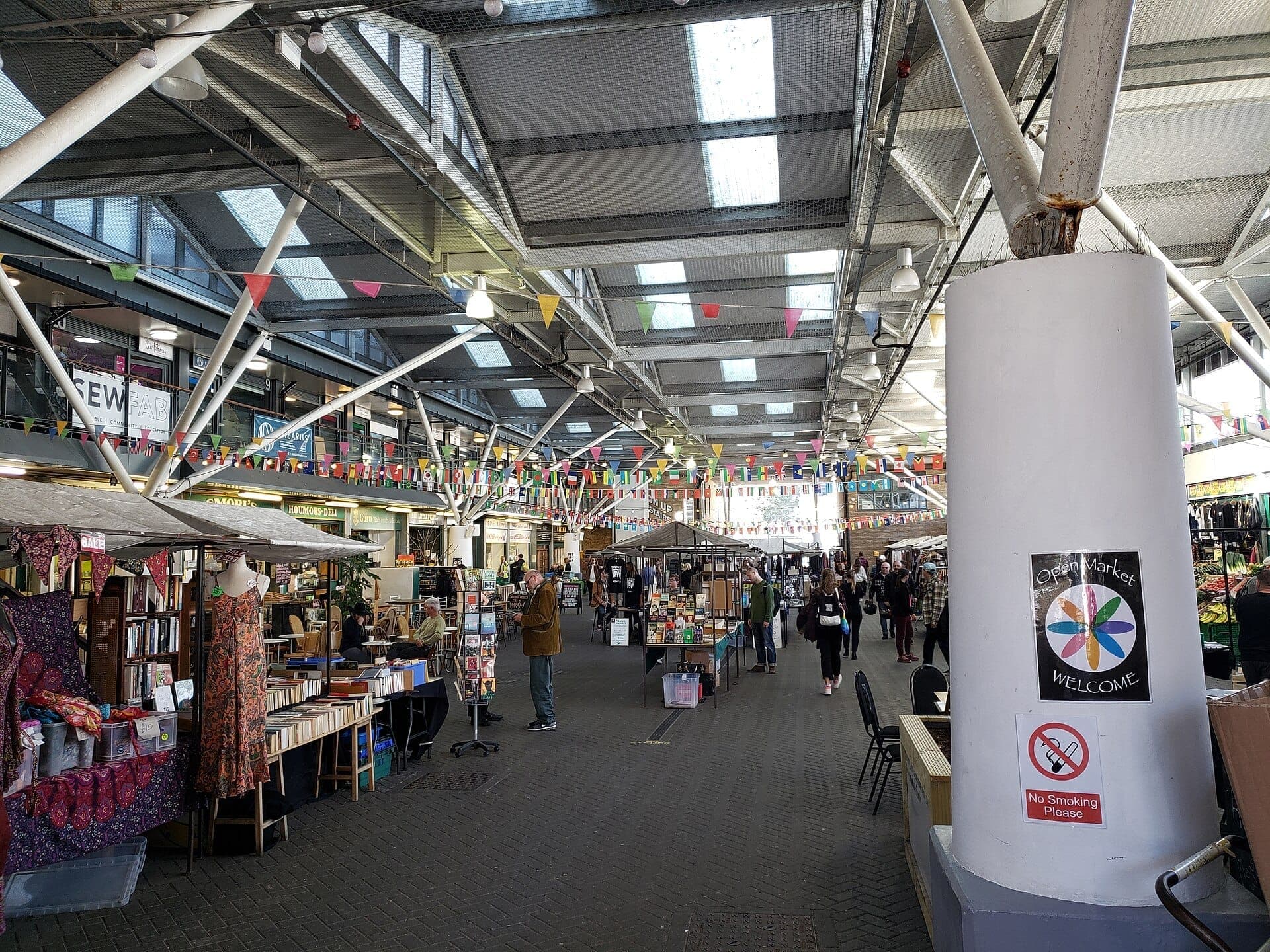 Brighton Open Market với mái che, dây cờ đầy màu sắc, các gian hàng độc lập, người mua sắm xem sách và hàng thủ công trong không gian trong nhà sôi động.