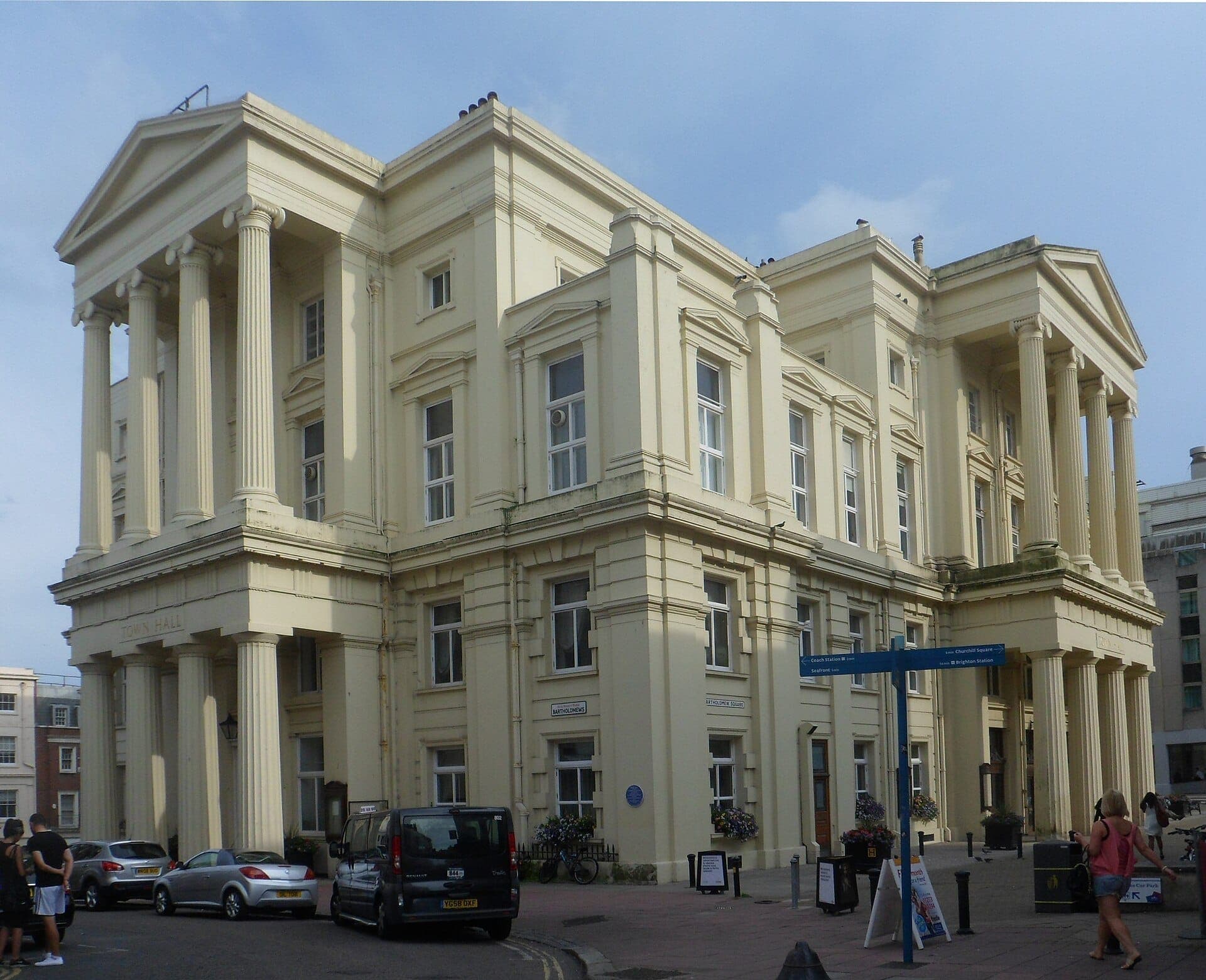 Mặt ngoài của Brighton Town Hall với những cột theo phong cách tân cổ điển hoành tráng, mặt tiền màu vàng nhạt, xe ô tô đậu và người đi trên đường dưới bầu trời quang đãng.