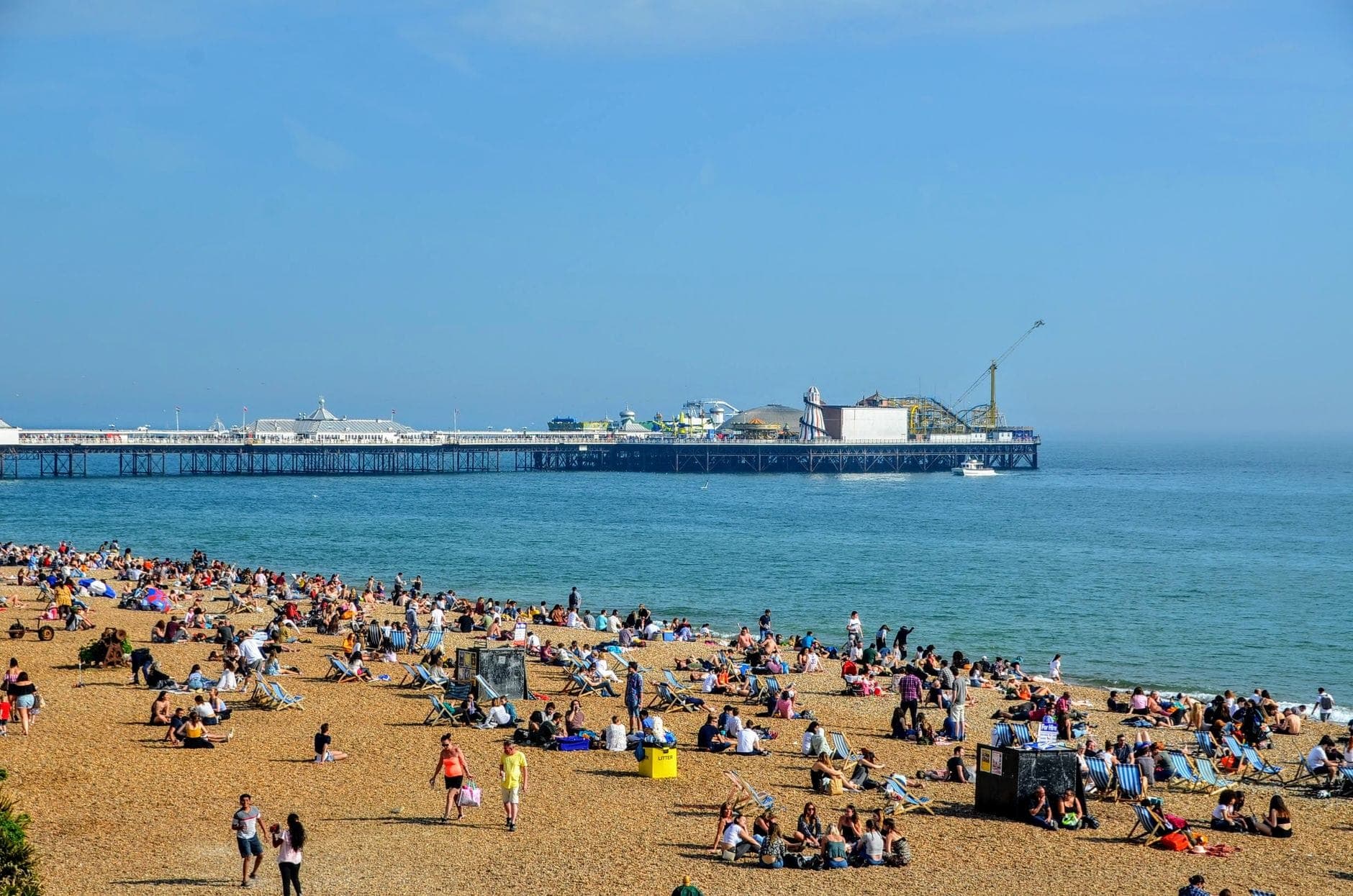 Khung cảnh sống động của bãi biển Brighton ngập tràn nắng, đông người thư giãn, phía xa là cầu tàu Brighton và biển xanh.