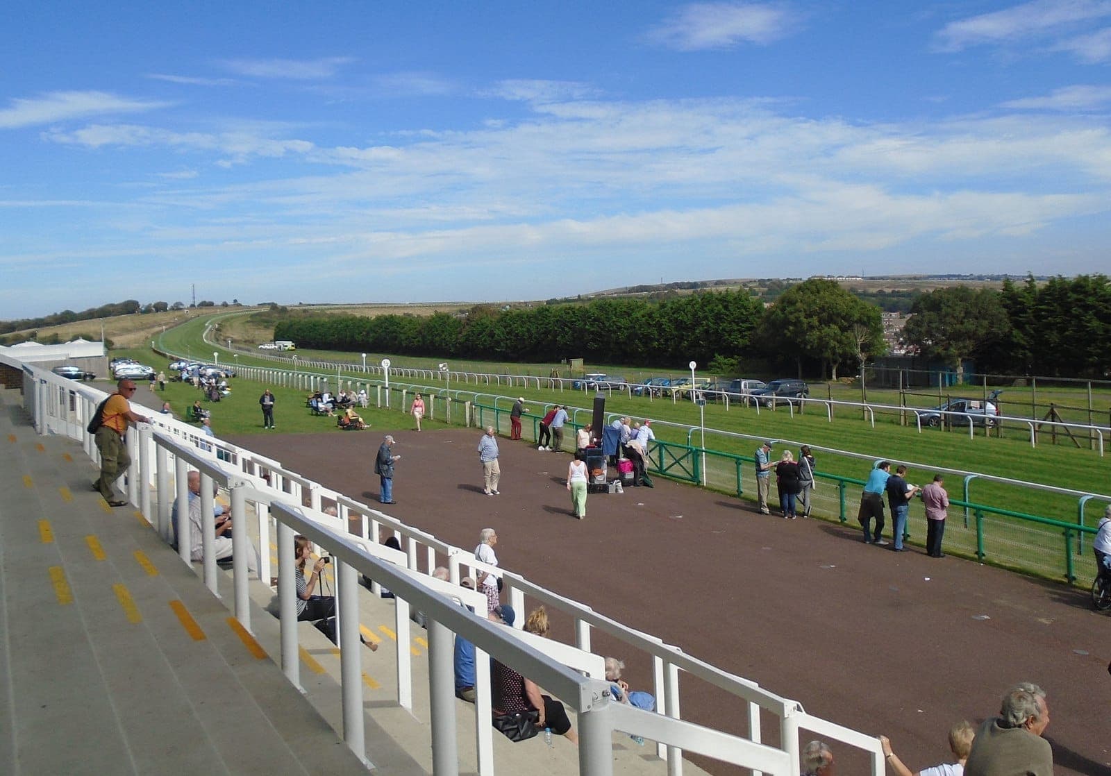 Khán giả xem từ khán đài tại Brighton Racecourse, nhìn ra đường đua xanh mướt, những ngọn đồi xa xa và các xe đậu trong một ngày nắng đẹp.