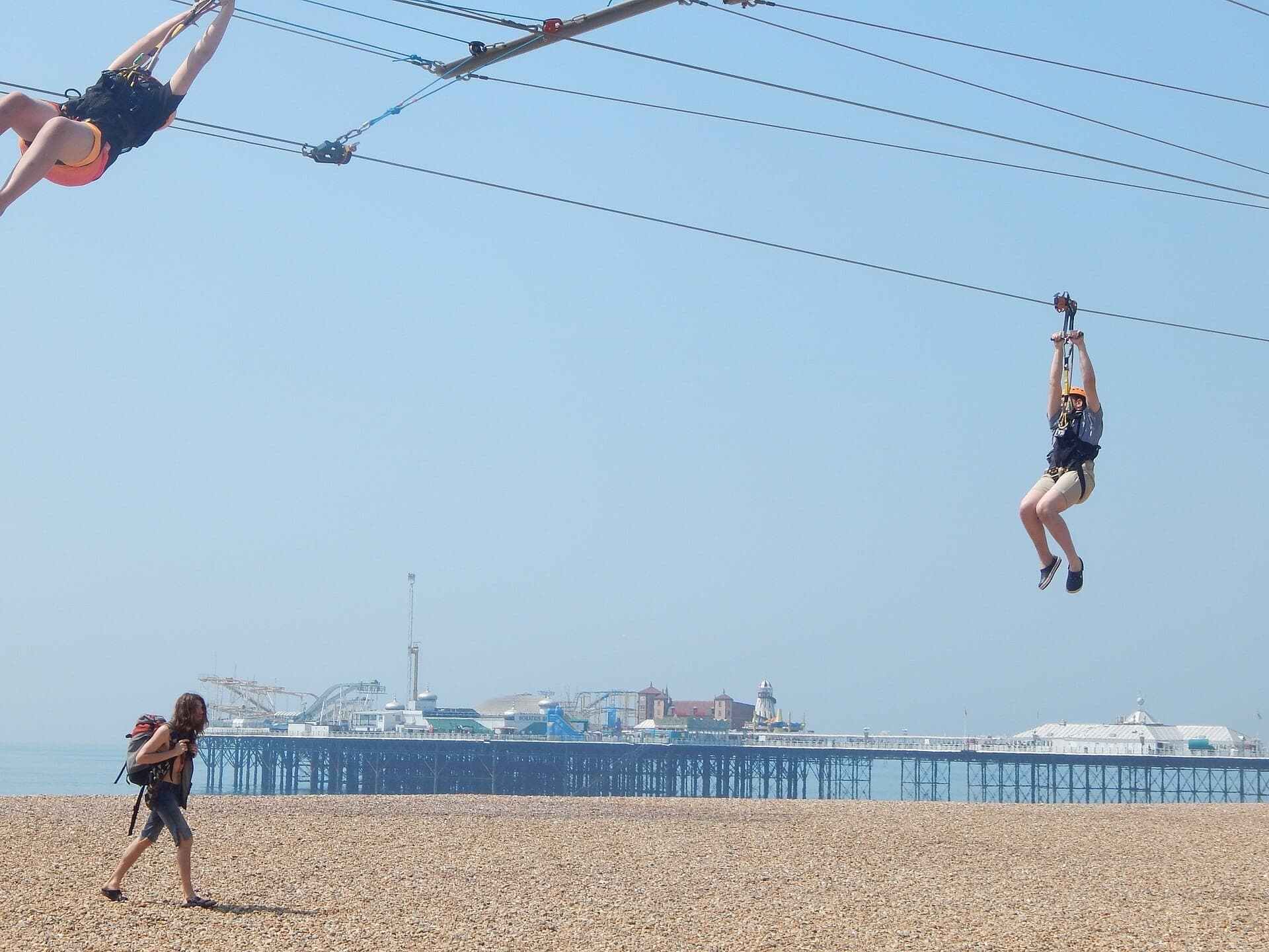 Hai người trượt Brighton Zip wire phía trên bãi biển sỏi, với Brighton Palace Pier hiện ra ở phía sau vào một ngày trời quang đãng.