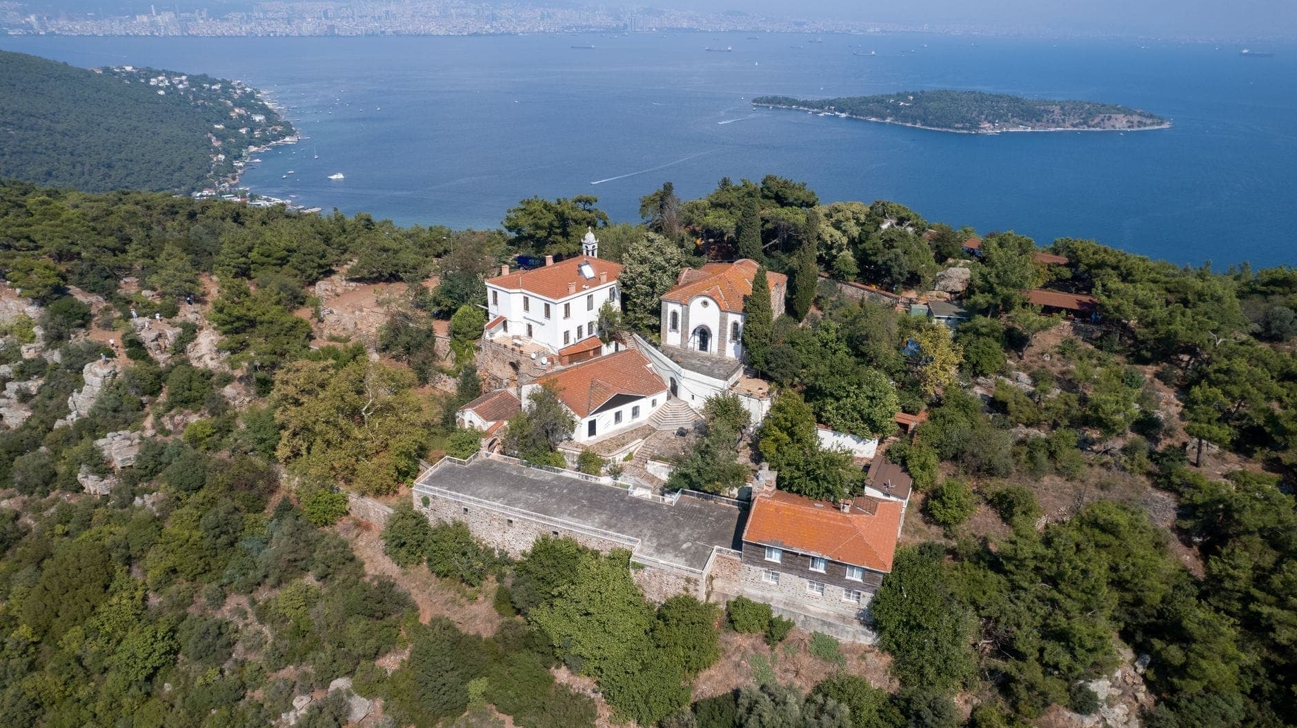 Luftaufnahme von Büyükada mit historischen Holzvillen, einer kleinen Kirche, üppigen Kiefernwäldern und dem tiefblauen Marmarameer – Istanbul ist im Hintergrund schemenhaft zu erkennen.