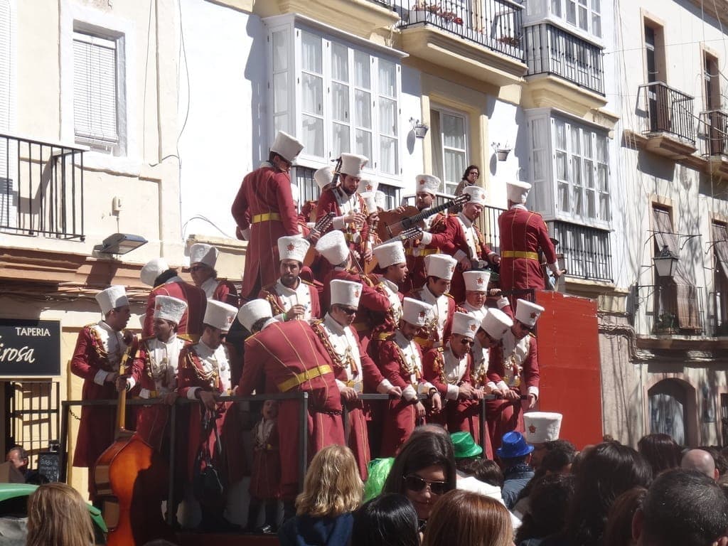 Một nhóm nhạc sĩ mặc trang phục đỏ trắng đồng bộ biểu diễn trên xe hoa trong lễ hội Carnaval Cádiz, với đám đông rực rỡ sắc màu và các tòa nhà lịch sử ở gần đó.