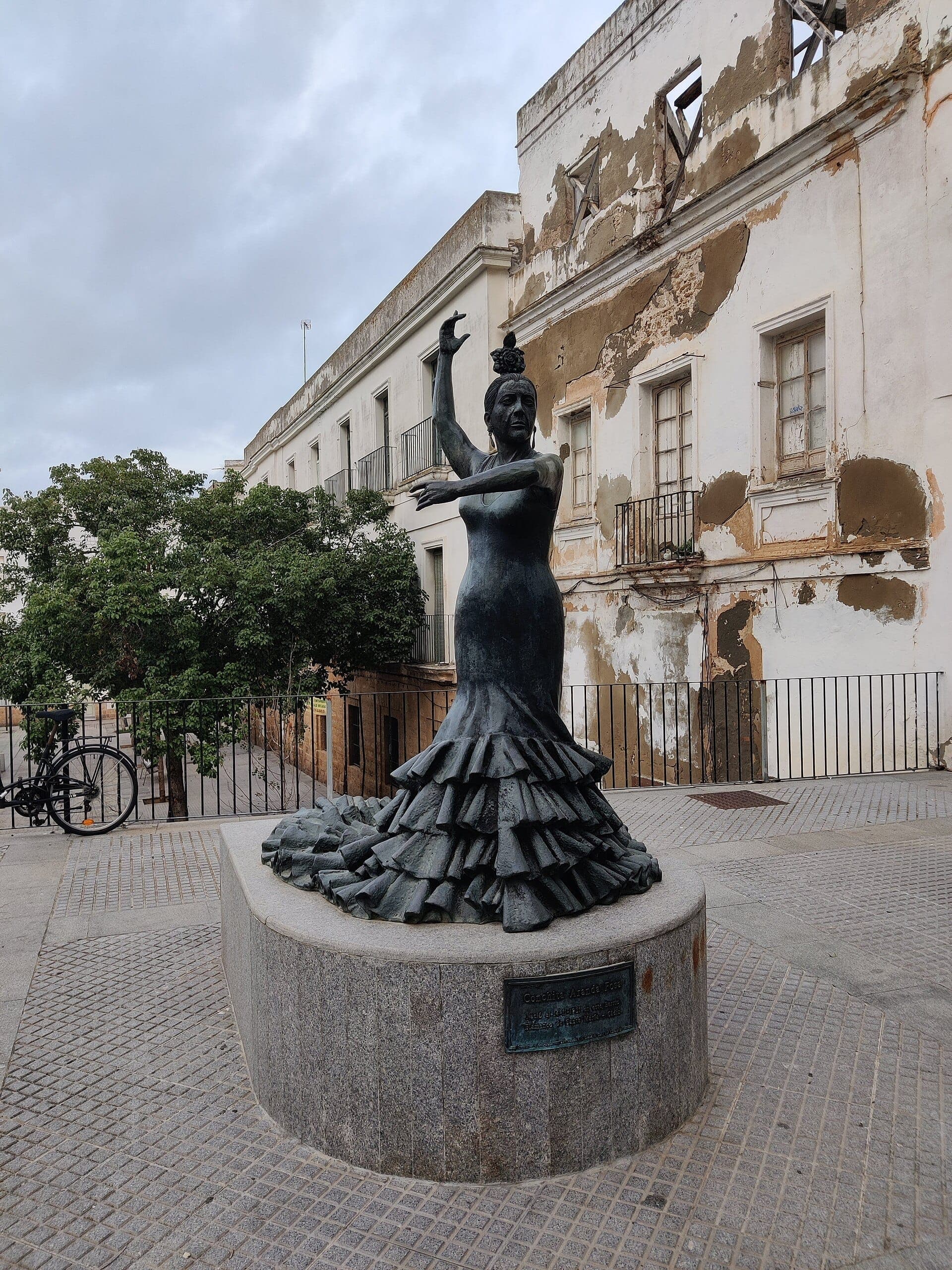 Tượng đồng vũ công flamenco tại Cádiz, Tây Ban Nha, đặt trước một tòa nhà cũ kỹ, xuống cấp với lớp sơn bong tróc và bầu trời nhiều mây.
