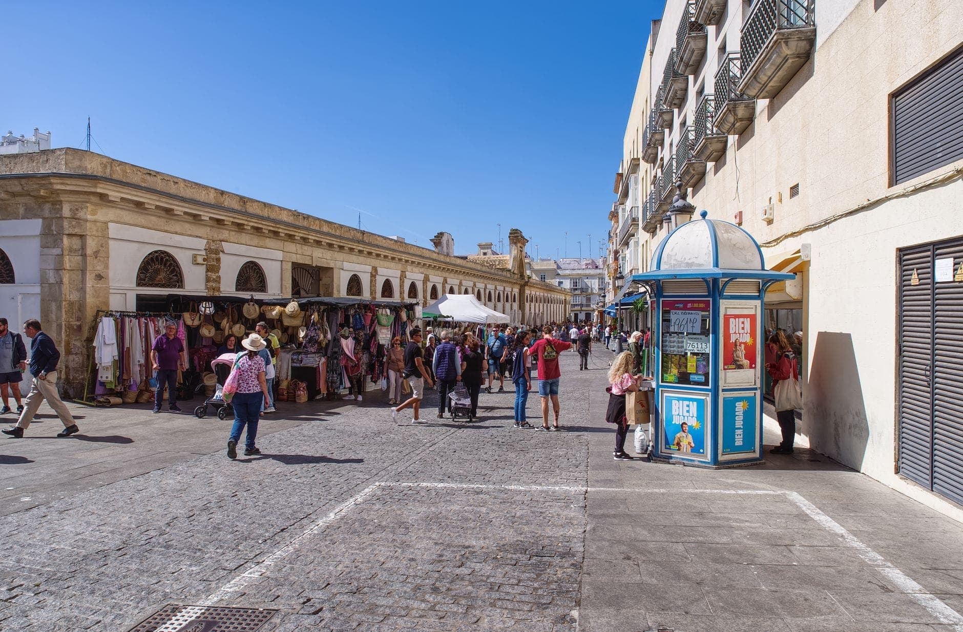 Cảnh phố chợ trời ở Cádiz với người dân mua sắm tại các sạp thực phẩm và thủ công dưới bầu trời xanh, không khí địa phương sống động.
