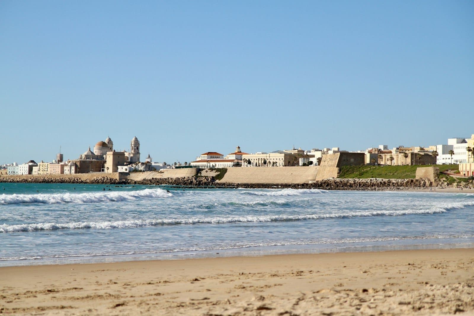 Toàn cảnh bãi biển Cádiz với sóng nhẹ, cát vàng óng và đường chân trời cổ kính dưới bầu trời xanh mùa hè.