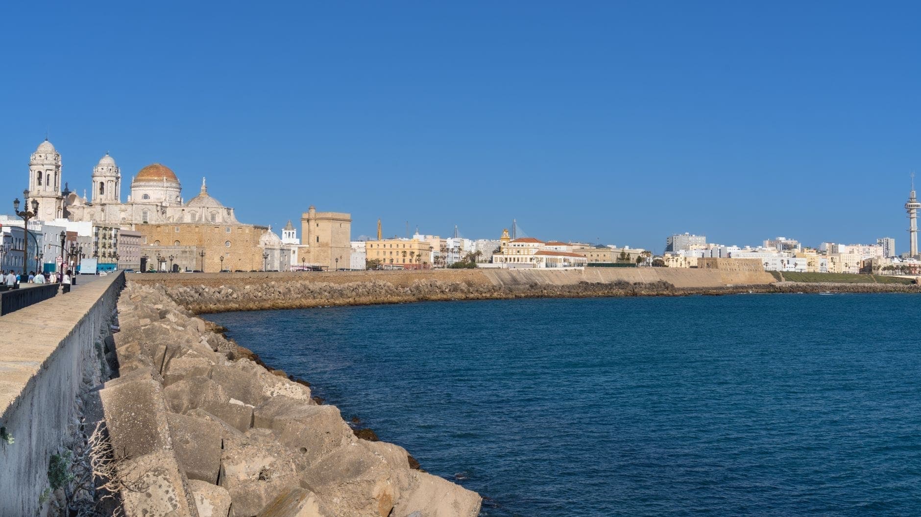 Toàn cảnh Cádiz nhìn từ bờ biển: mái vòm nhà thờ cổ, tường thành, những ngôi nhà và biển Đại Tây Dương xanh thẳm dưới bầu trời quang đãng.