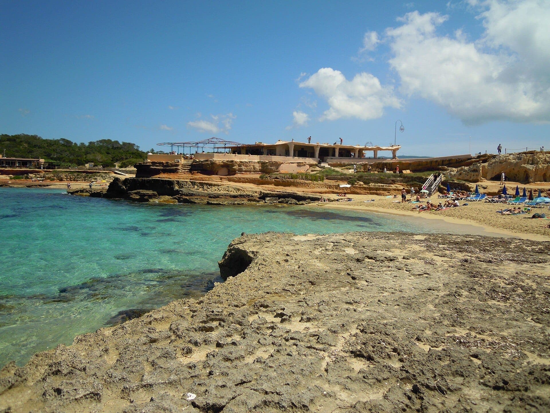 View of Cala Comte beach in Ibiza with clear turquoise water, rocky shoreline, sandy coves, sunbathers, and a rustic shoreline restaurant.