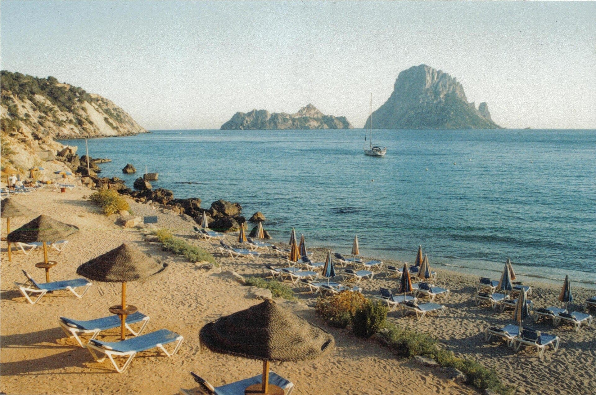 Cala d'Hort beach with rows of empty sun loungers and straw umbrellas facing the blue sea and rocky Es Vedrà island in the distance.