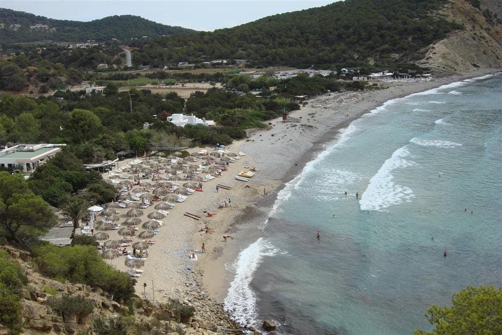 Aerial view of Cala Jondal beach with white pebbles, turquoise water, sun loungers, and beach clubs surrounded by green hills.