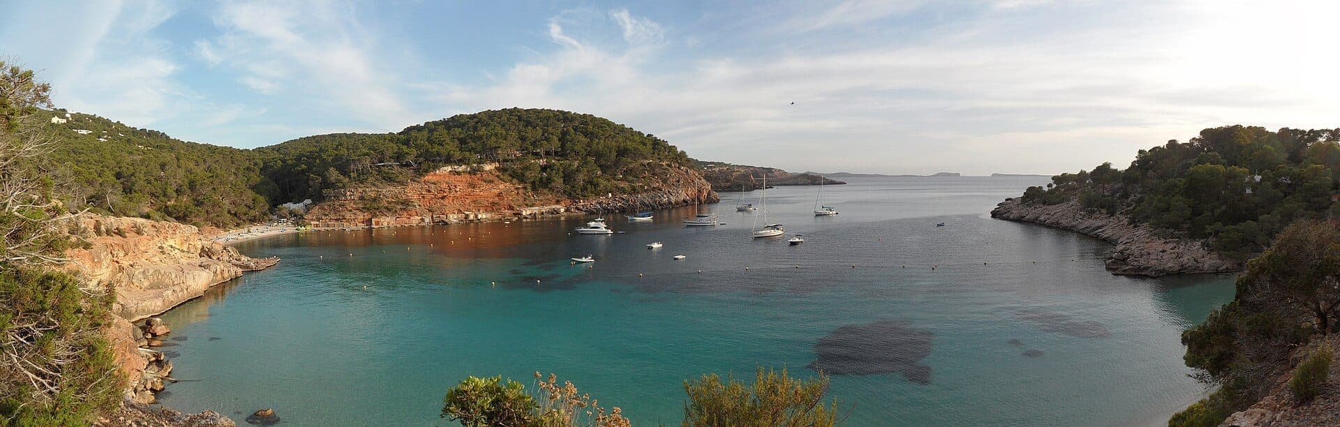 Panoramic view of Cala Salada and Cala Saladeta coves in Ibiza, featuring turquoise waters, anchored boats, rocky cliffs, and lush green hills.