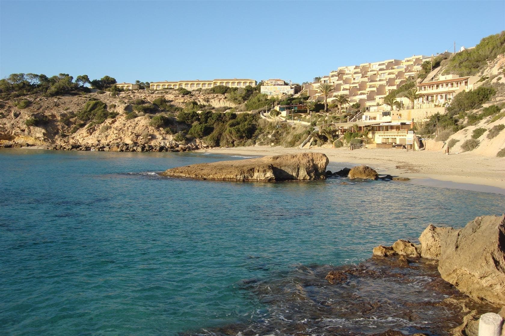 Clear turquoise waters and white sand at Cala Tarida, with rocky outcrops and hillside resorts on Ibiza’s scenic west coast.