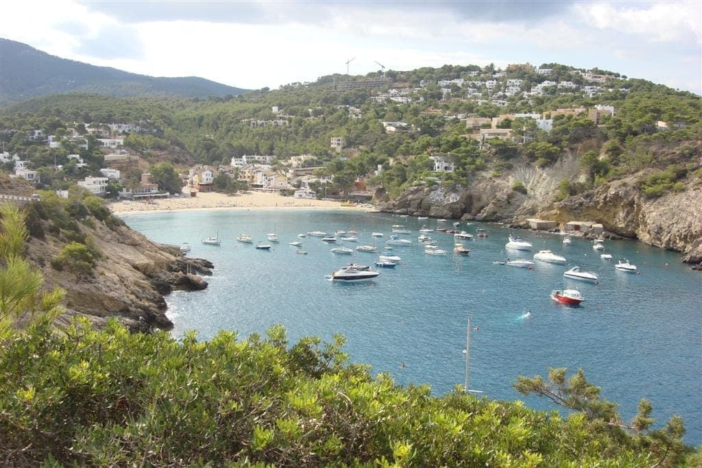 View of Cala Vadella cove in Ibiza showing turquoise water, anchored boats, sandy beach, and white hillside villas surrounded by green hills.