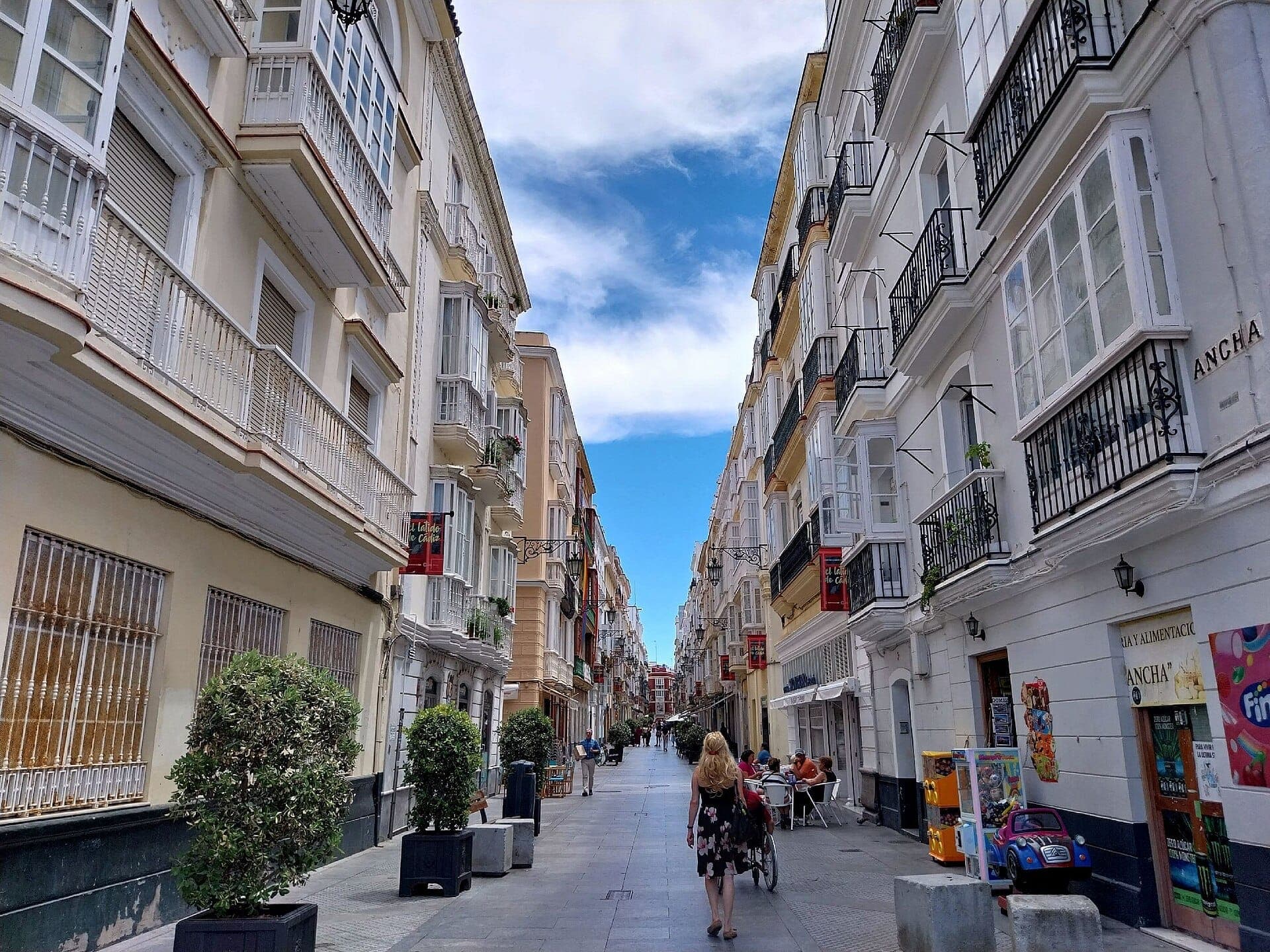 Cảnh rộng của Calle Ancha ở Cádiz với những tòa nhà lịch sử, cây trồng trong chậu, ban công mở, quán cà phê ngoài trời và người dân dạo bộ dưới bầu trời trong sáng.