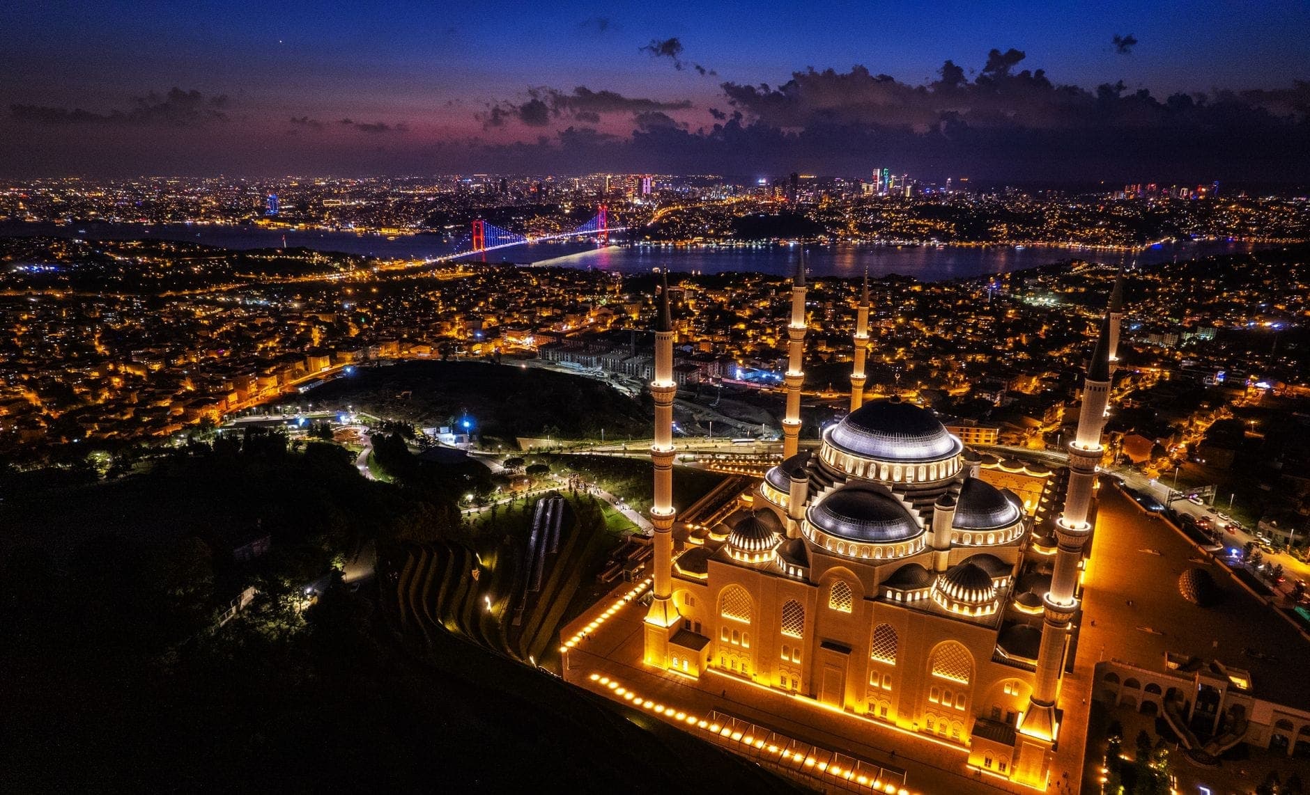 Luftaufnahme der Çamlıca-Moschee in der Abenddämmerung, beleuchtet vor der Skyline Istanbuls und der Bosporus-Brücke im Hintergrund.