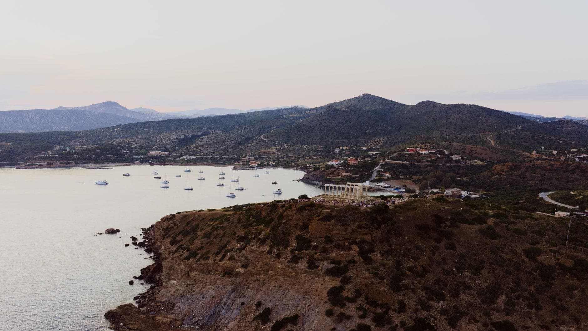 A dramatic aerial view of Cape Sounion showing the Temple of Poseidon perched on a cliff above the Aegean Sea, with anchored boats and rolling hills in the background.