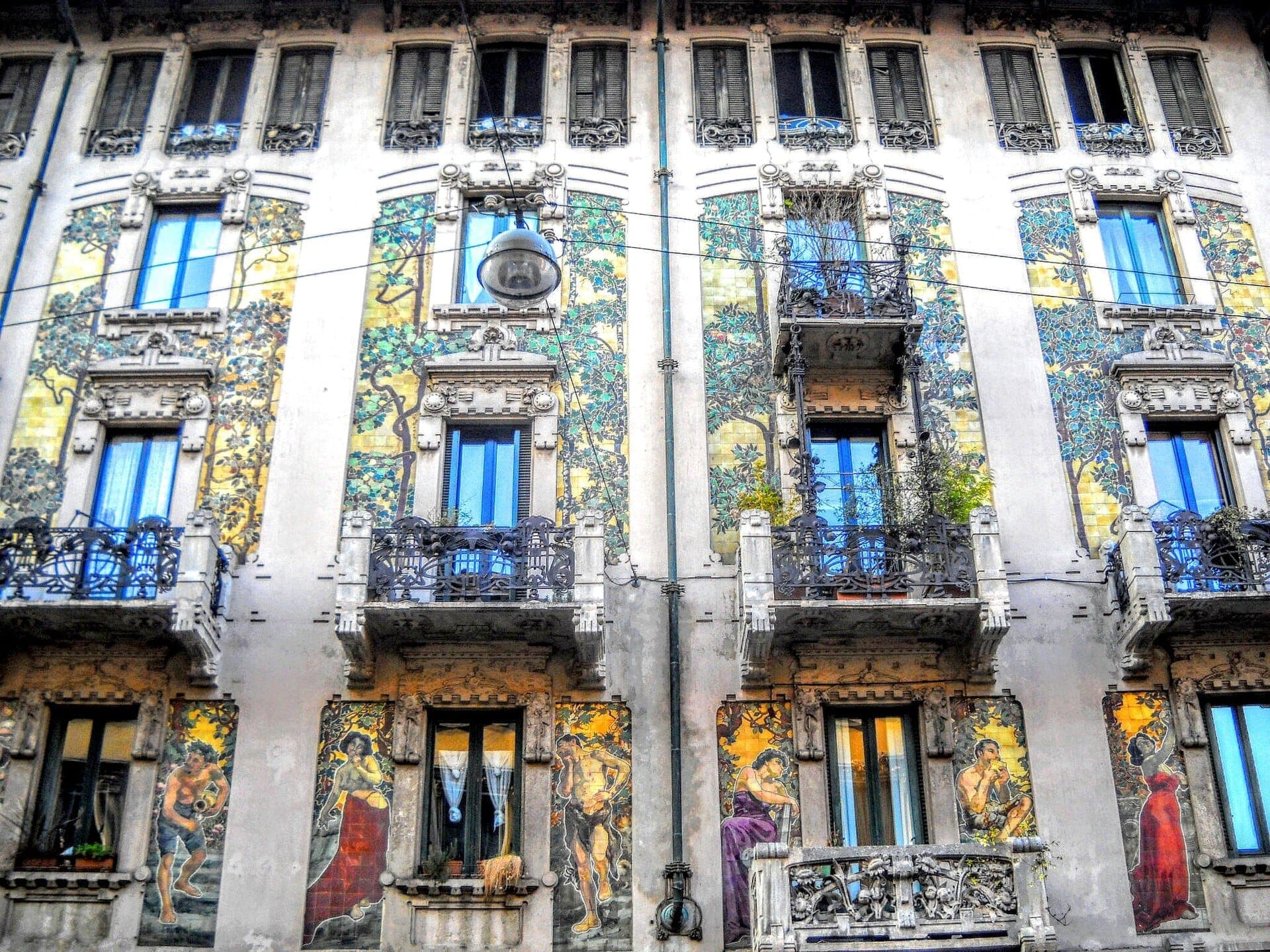 Front view of Casa Galimberti’s Liberty style façade, showcasing intricate wrought iron balconies and vibrant ceramic panels with floral and human figures.