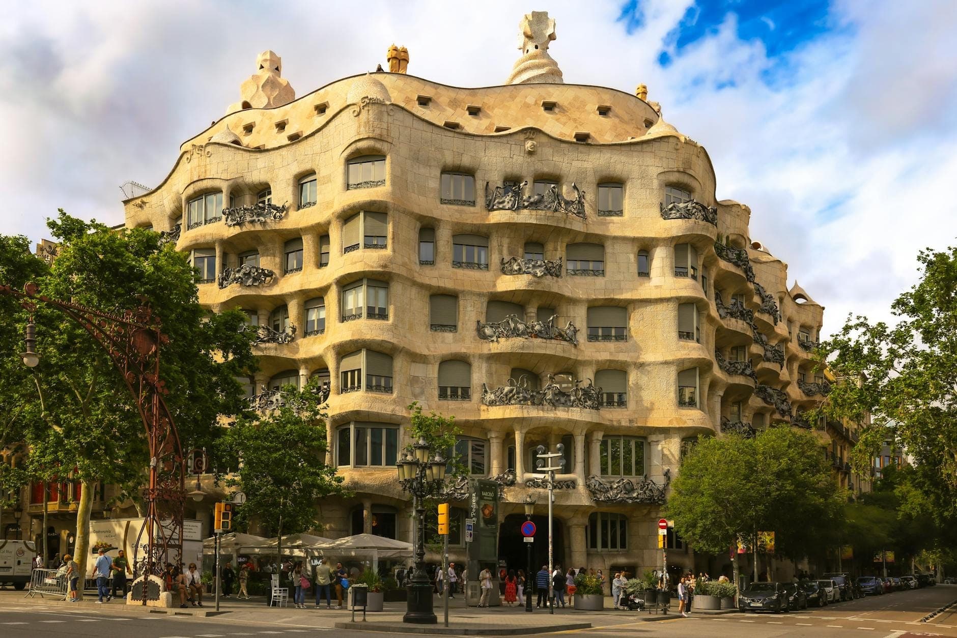 Toàn cảnh mặt tiền đá gợn sóng nổi tiếng của Casa Milà với những ban công sắt rèn tinh xảo dưới bầu trời xanh có mây, nhộn nhịp du khách và đời sống đường phố Barcelona.