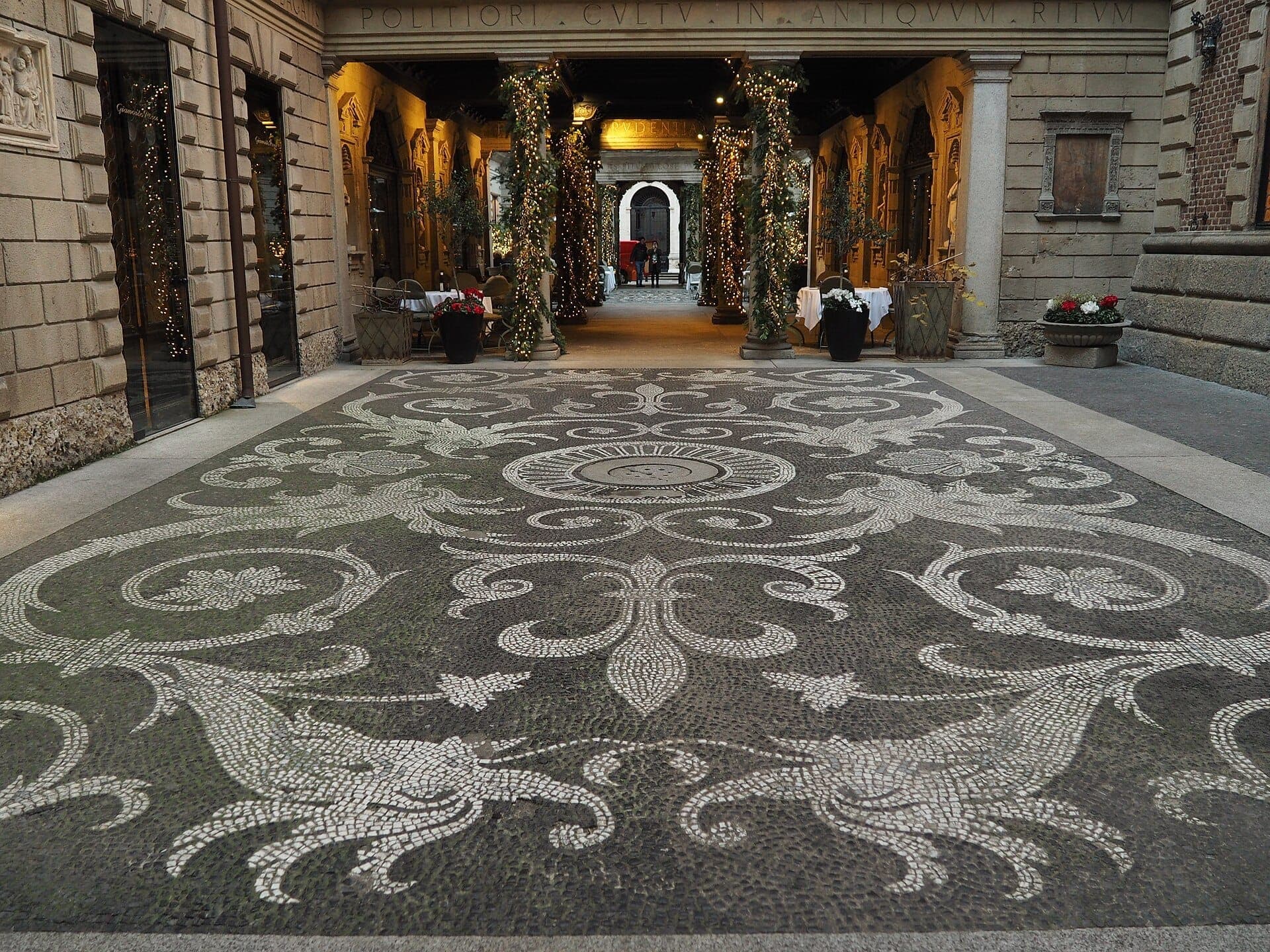 Elegant courtyard entrance of Casa Museo Bagatti Valsecchi in Milan with intricate mosaic floor and illuminated arches, surrounded by historic stone buildings.