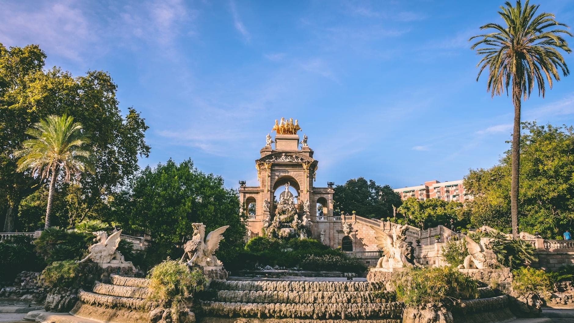 Toàn cảnh đài phun nước Cascada Monumental trong Parc de la Ciutadella, Barcelona, với cây xanh tươi tốt, những bức tượng cầu kỳ, mặt hồ xanh ngọc và tượng vàng dưới bầu trời xanh.