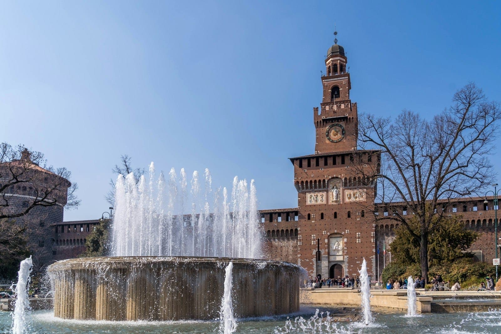 Castello Sforzesco in Milan with its brick facade and clock tower rises behind a large circular fountain on a sunny day.
