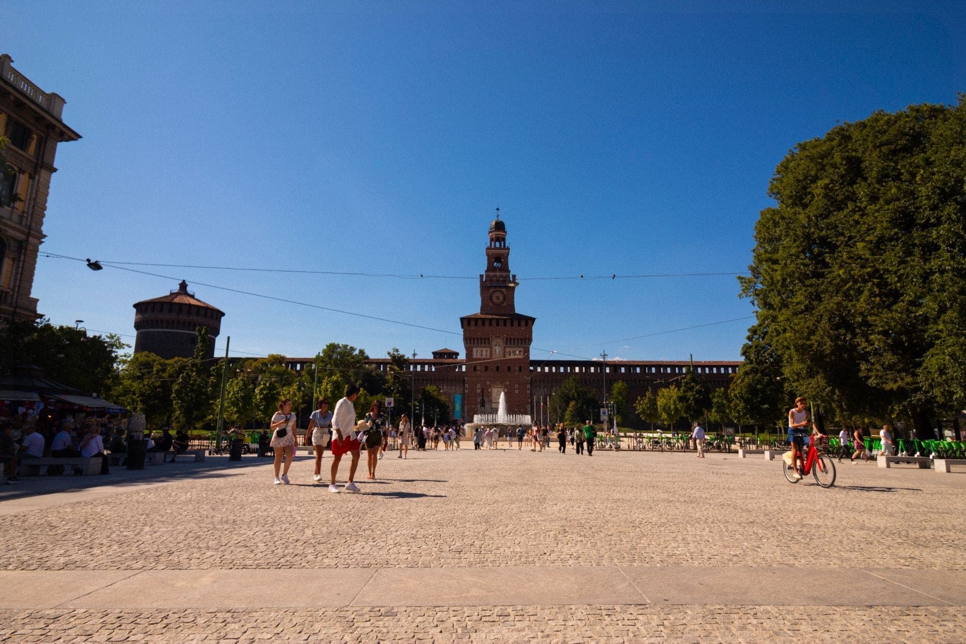 Wide view of Castello Sforzesco’s main facade on a sunny day, with visitors walking and cycling along the open cobbled square, framed by lush green trees.