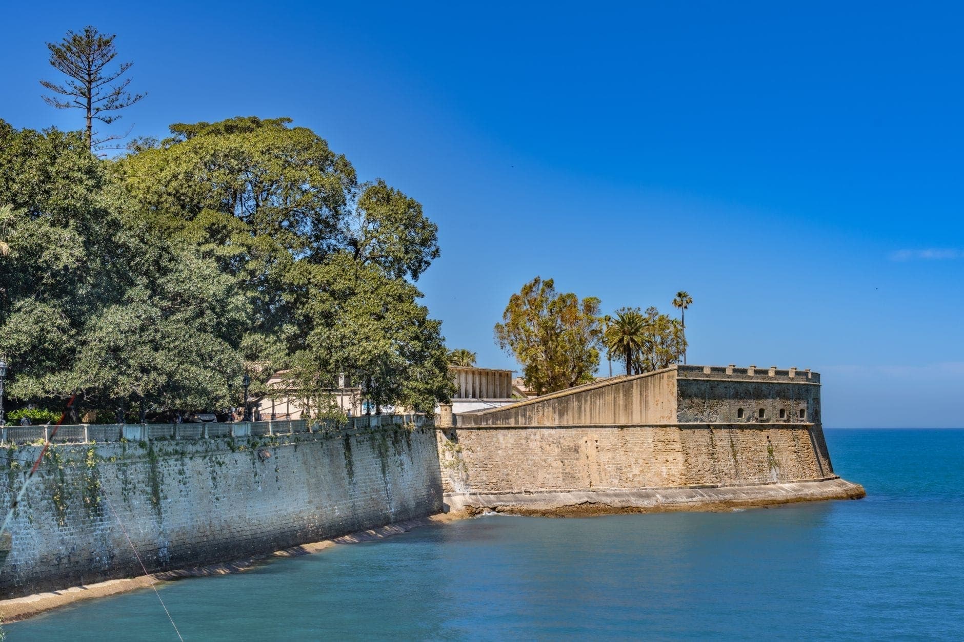 Tường đá của Castillo de Santa Catalina vươn ra biển xanh dưới nắng, xung quanh là cây xanh và bầu trời trong ở Cádiz, Tây Ban Nha.