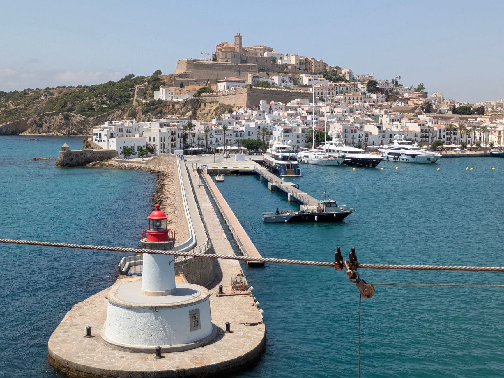 View of the Castle of Ibiza perched atop Dalt Vila with whitewashed buildings, a marina with yachts, and a lighthouse in the foreground.